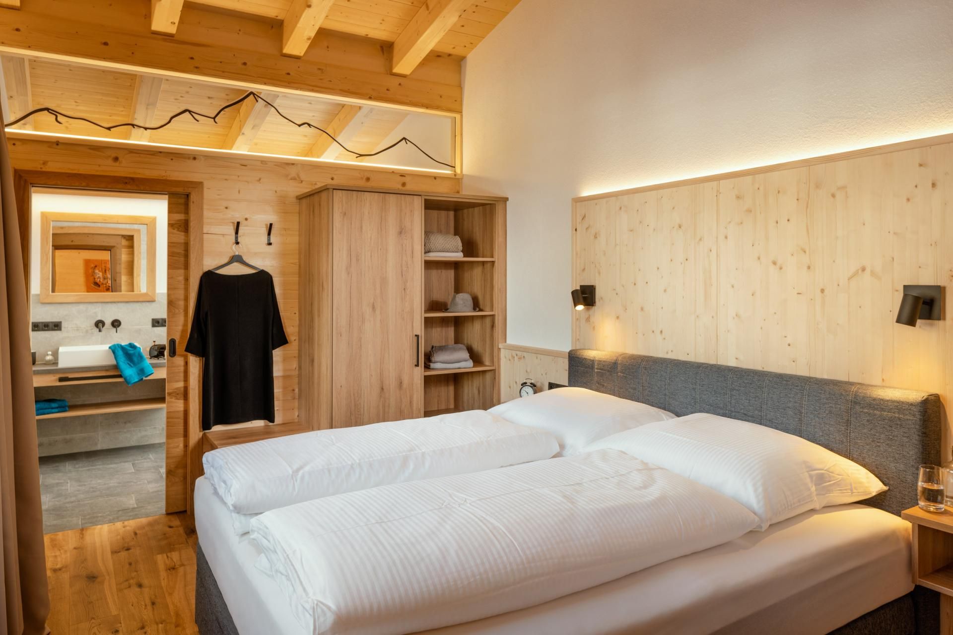 A modern bedroom with bright wood decor and a large double bed. In the background, a bathroom with a wooden shelf is visible.