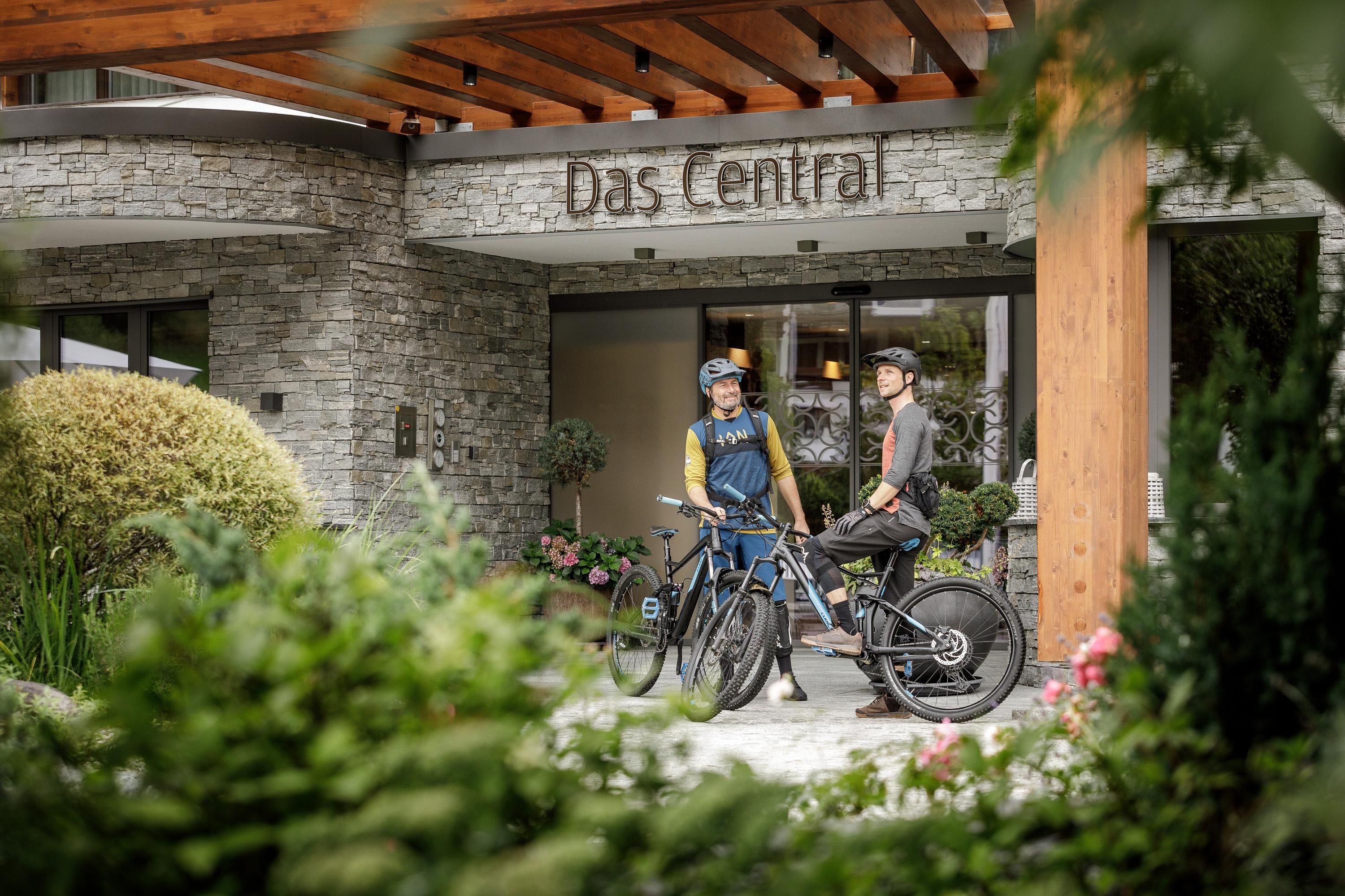 Two cyclists are standing in front of the hotel "Das Central." The surroundings are green and inviting, with modern architecture in the background.