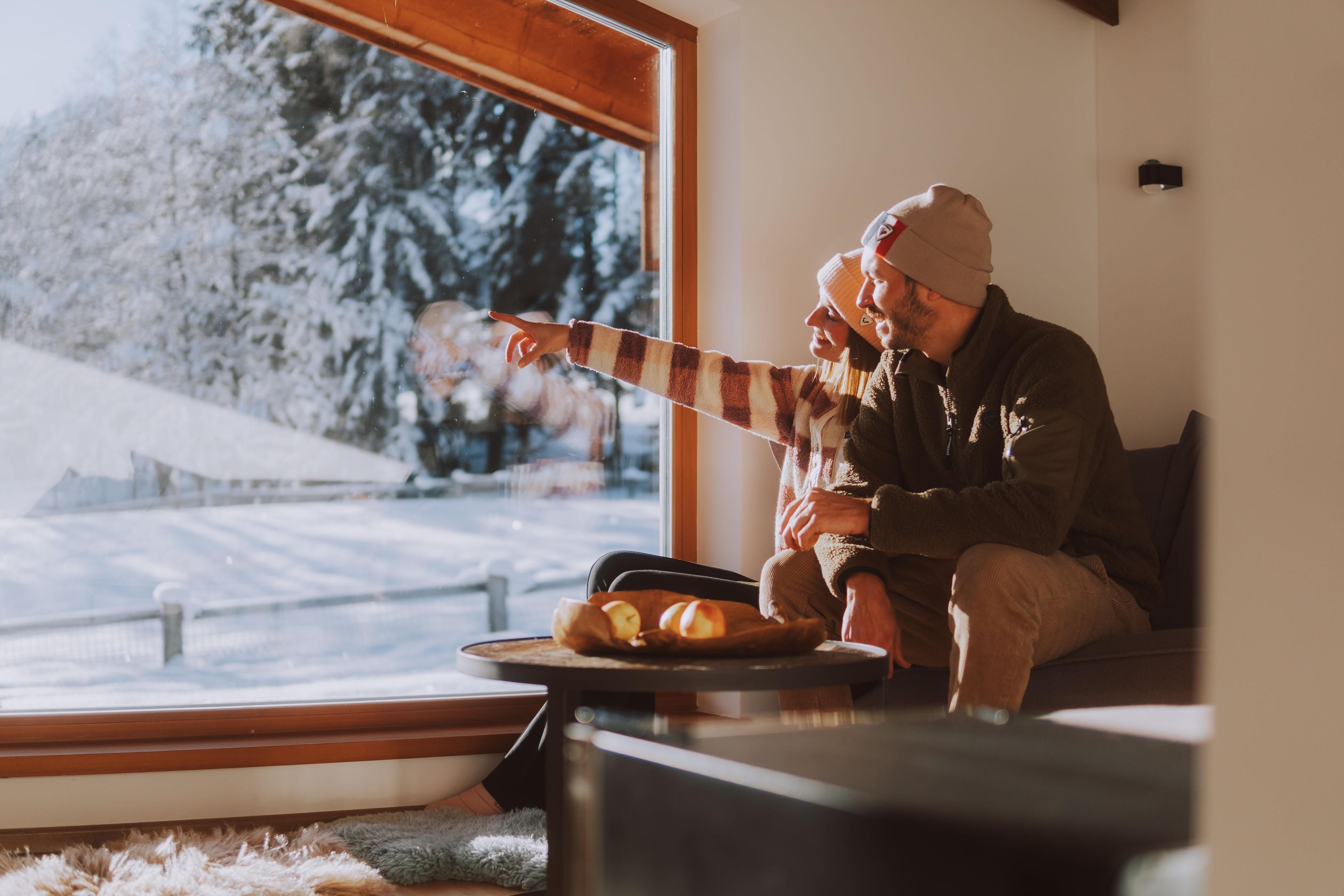 A couple sits comfortably in the living room and looks out the window. Outside, snow lies on the ground and the landscape is wintry.