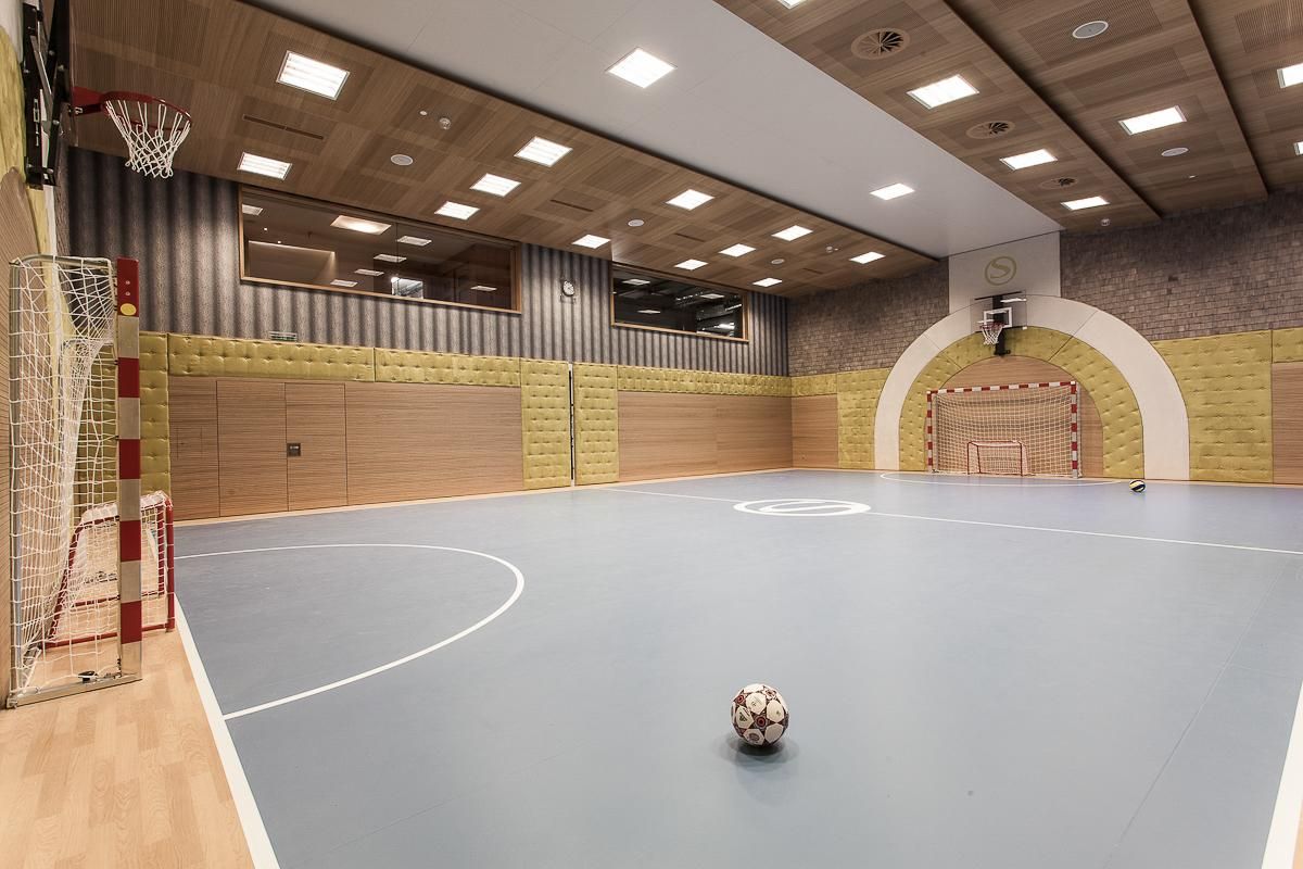 A gymnasium with a blue floor and a handball goal. A soccer ball is in the middle of the room.