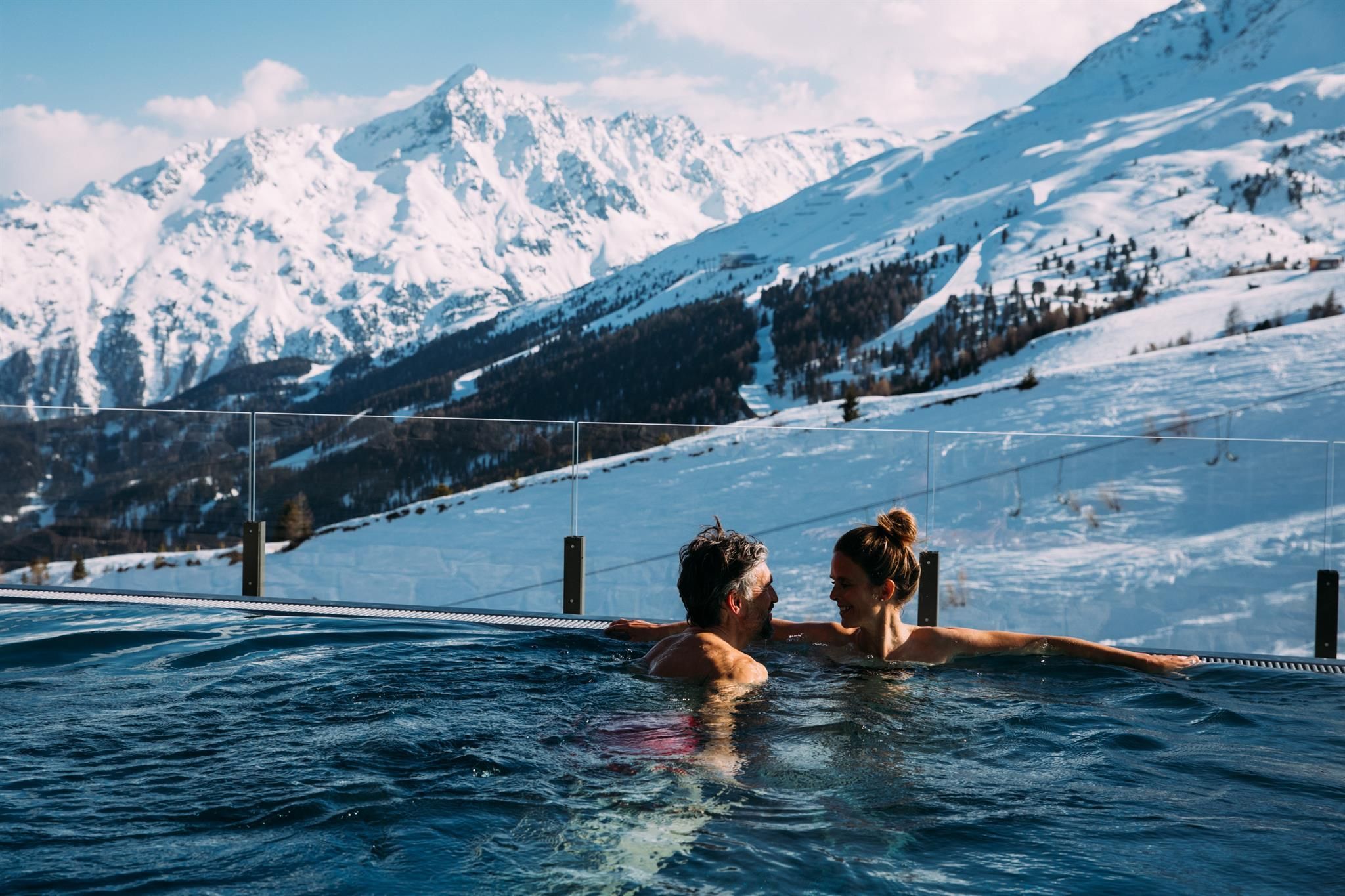 A couple relaxes in an outdoor pool with snow-covered mountains in the background. The winter landscape provides a breathtaking backdrop for a romantic getaway.