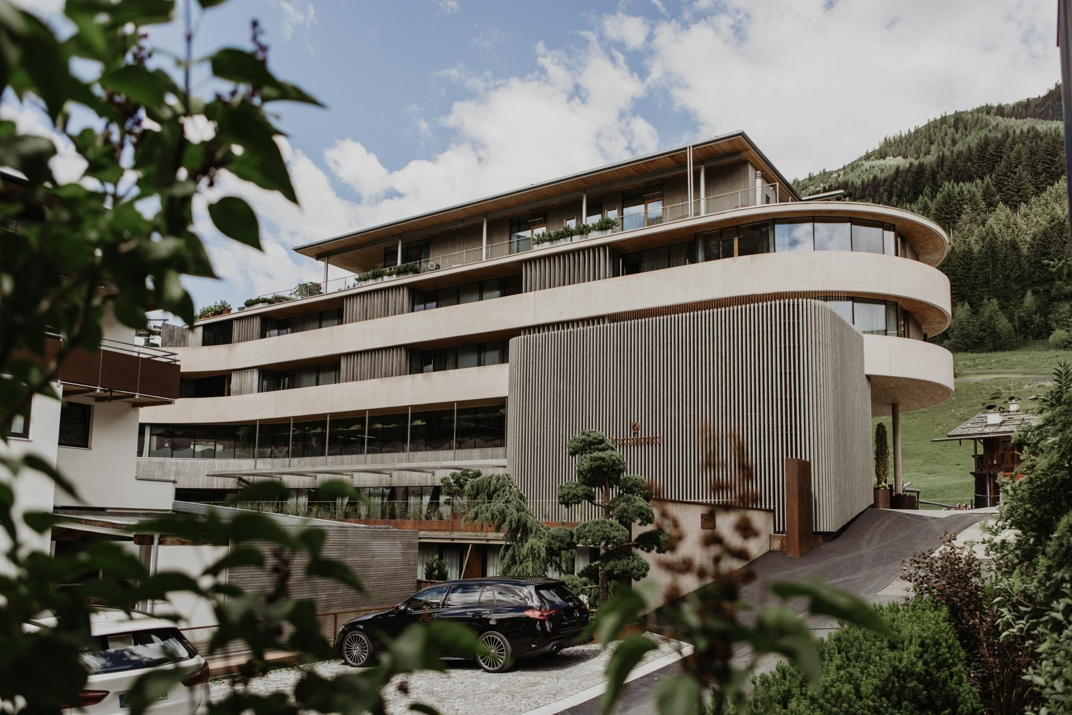 A modern building with a curved facade and large windows. It is surrounded by green hills and appears inviting.