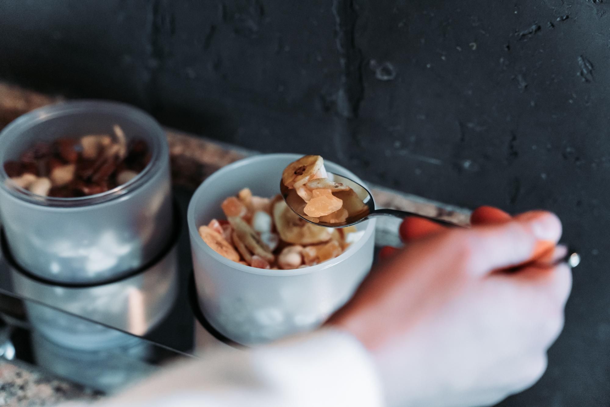 A hand is holding a spoon with nuts and a small bowl. In the background, other containers with nuts are visible.