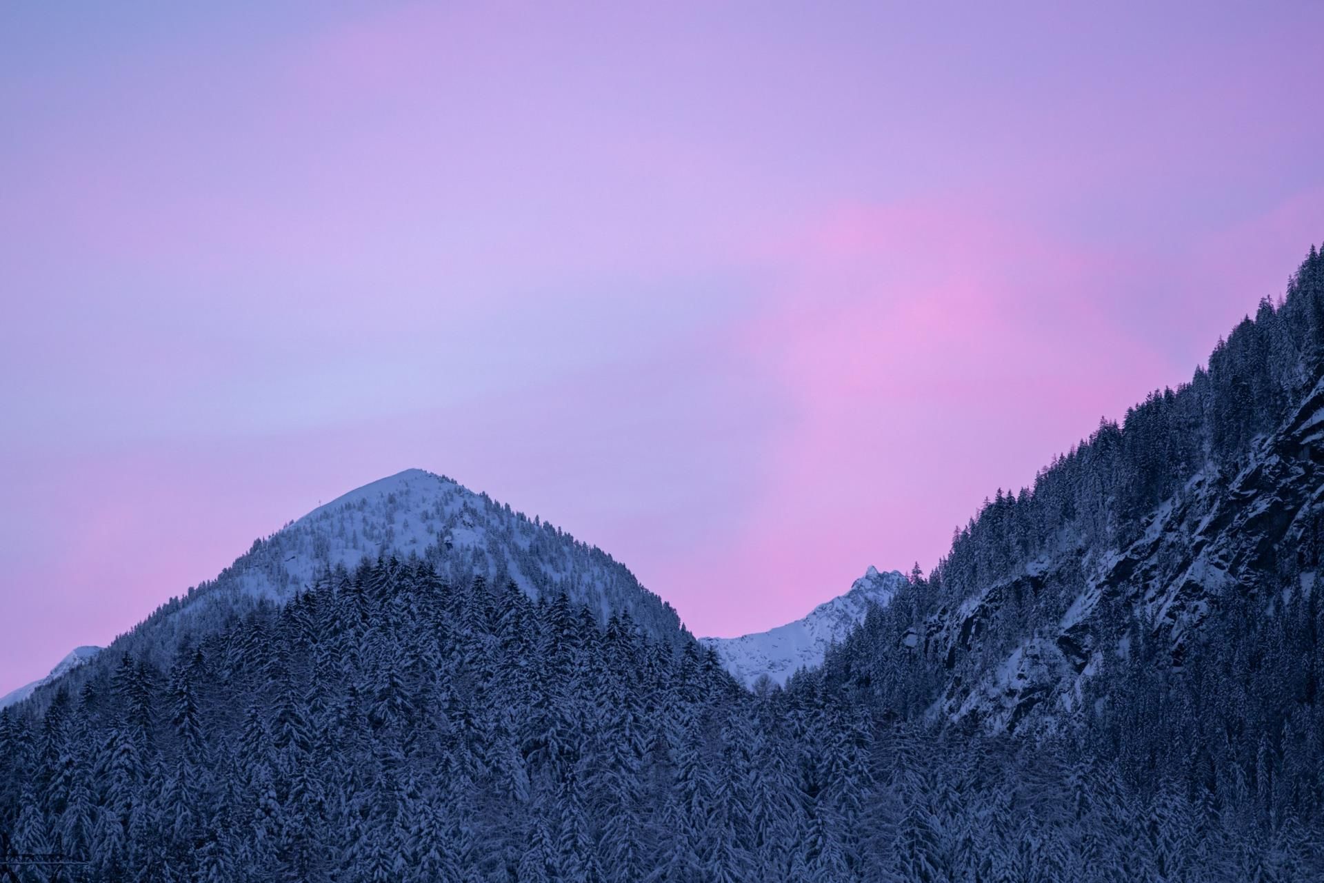 A wintry mountain landscape with snow-covered peaks. The sky radiates in gentle shades of pink and purple.