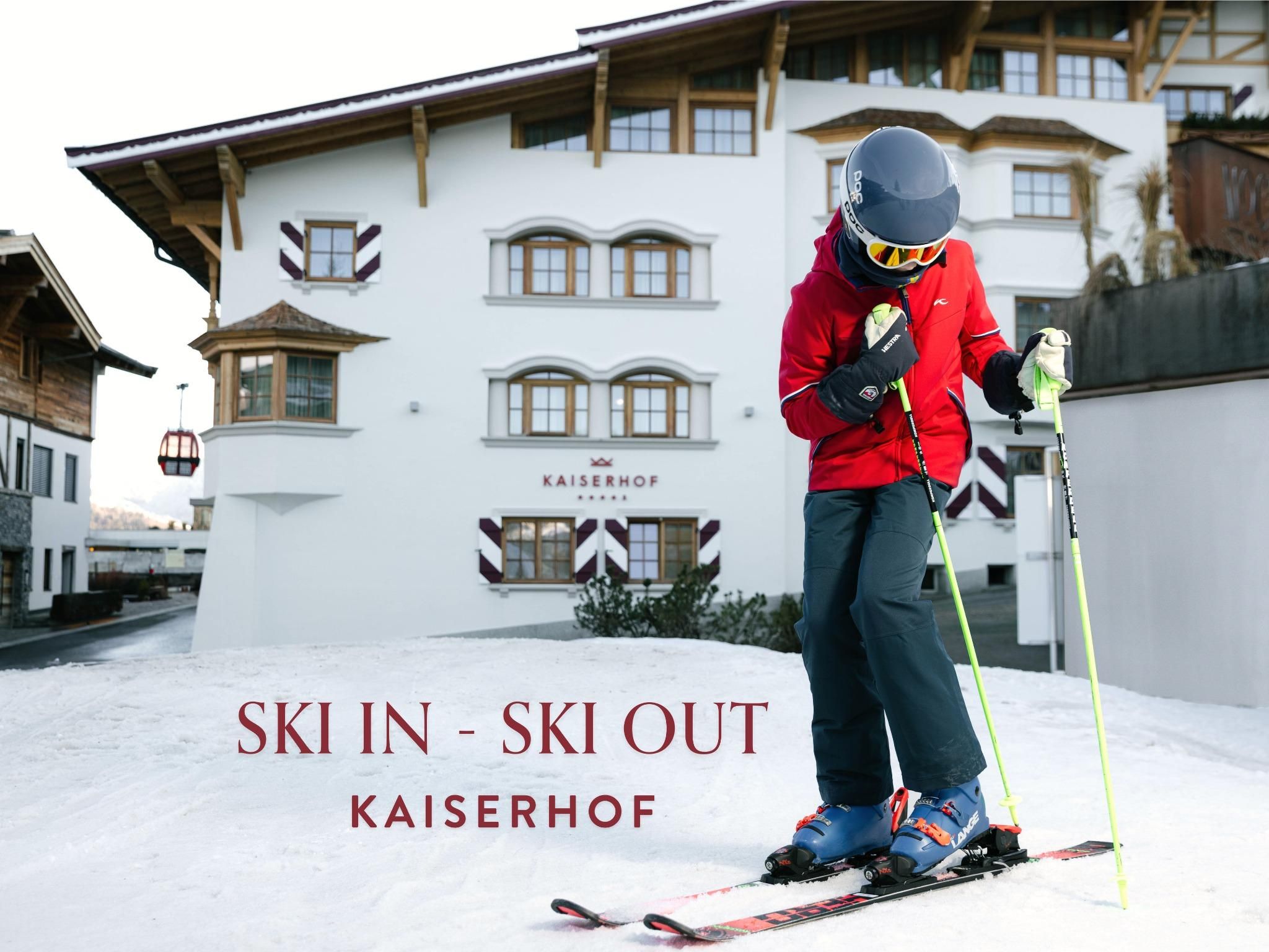 A skier stands on snow in front of the Hotel Kaiserhof. In the background, traditional Alpine architecture and the inscription "SKI IN - SKI OUT" can be seen.