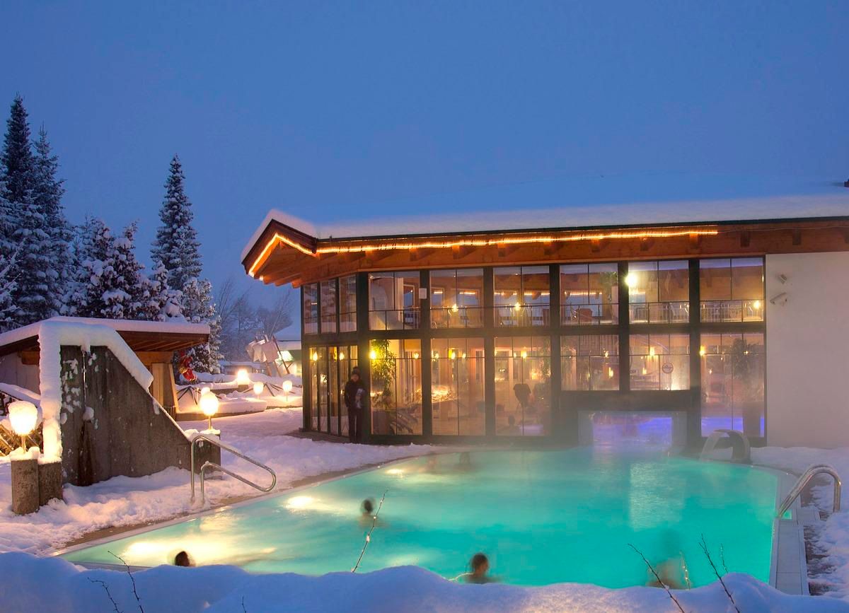 A modern spa building with large windows in a snowy landscape. In the foreground, there is a heated outdoor pool, surrounded by snow-covered trees.