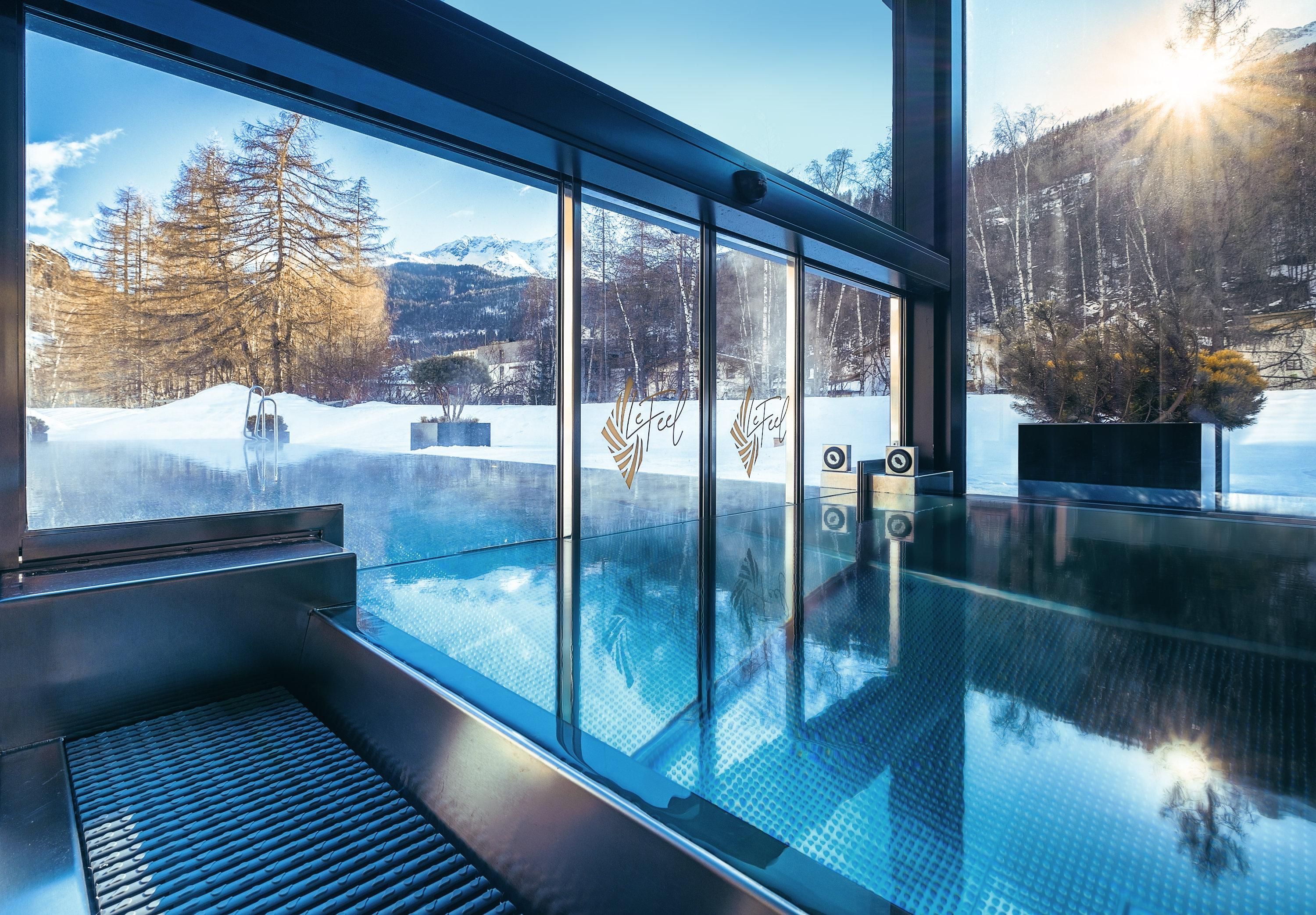 A modern swimming pool with large windows offers a view of snow-covered mountains and trees. The sun shines warmly into the room.