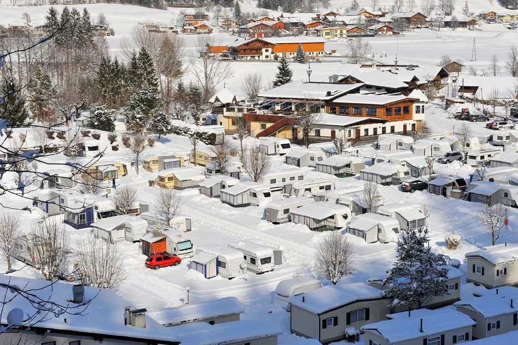 A snow-covered campsite landscape with caravans and surrounding trees. In the background, wooden buildings and snow-covered mountains can be seen.