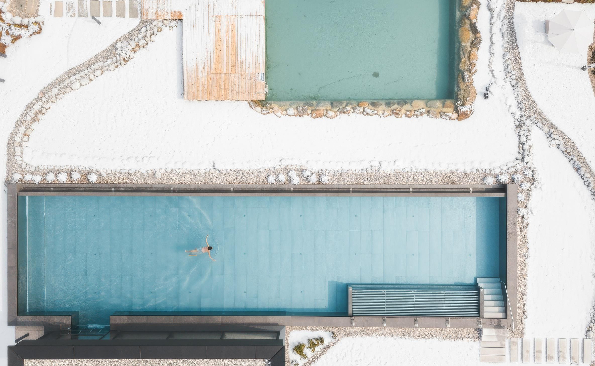 A narrow pool in a snow-covered setting, where a person is swimming. In the background, two more bodies of water are visible.