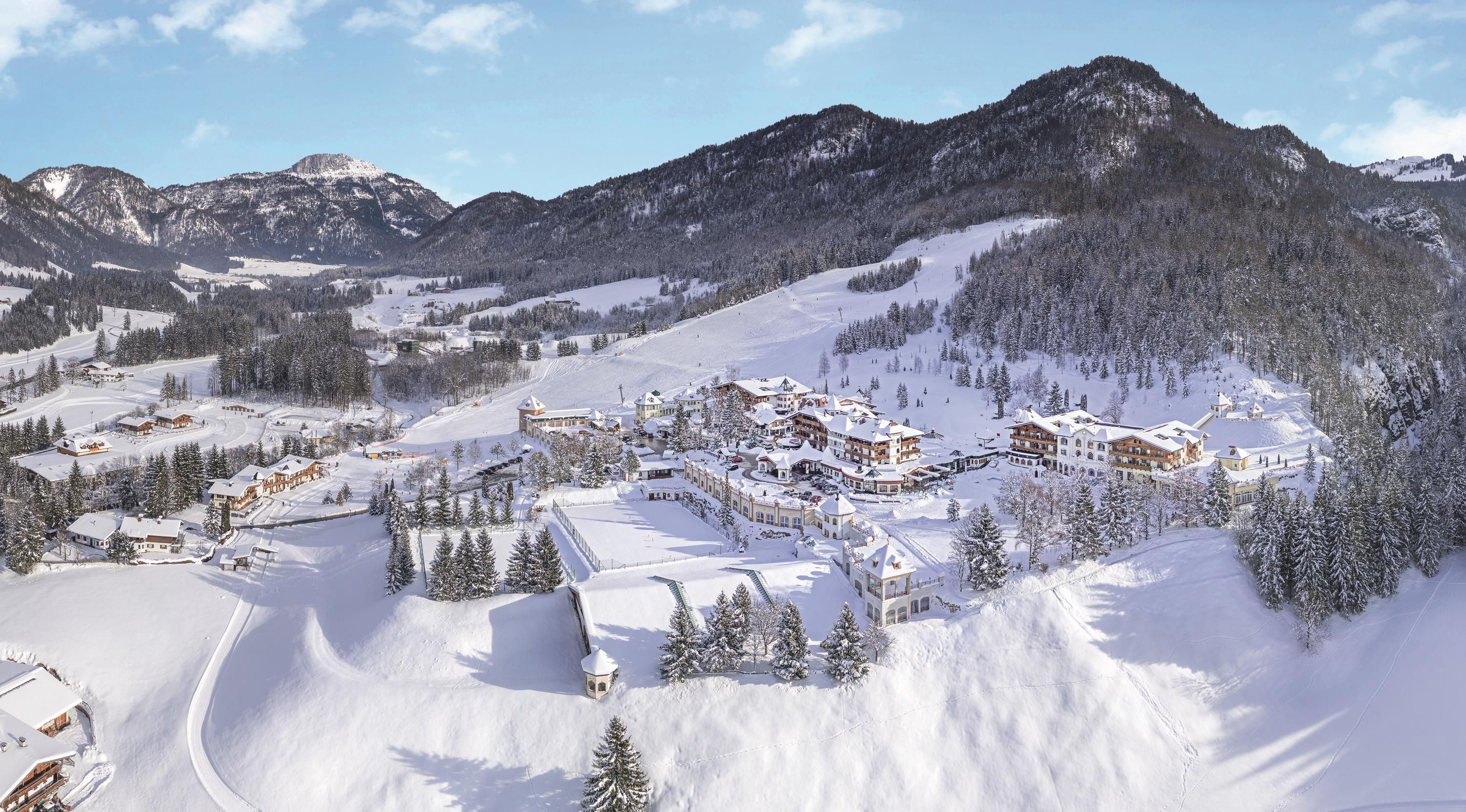A picturesque winter landscape with snow-covered mountains and a cozy holiday resort. The trees are covered with snow, and the sky is clear and blue.