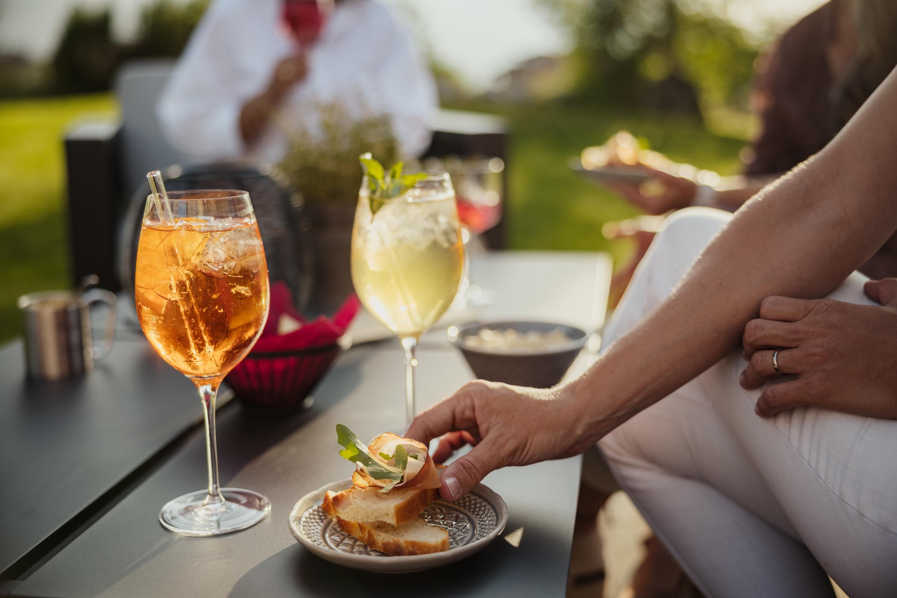 An outdoor table with two delicious cocktails and a person enjoying a snack. In the background, other people are relaxing.
