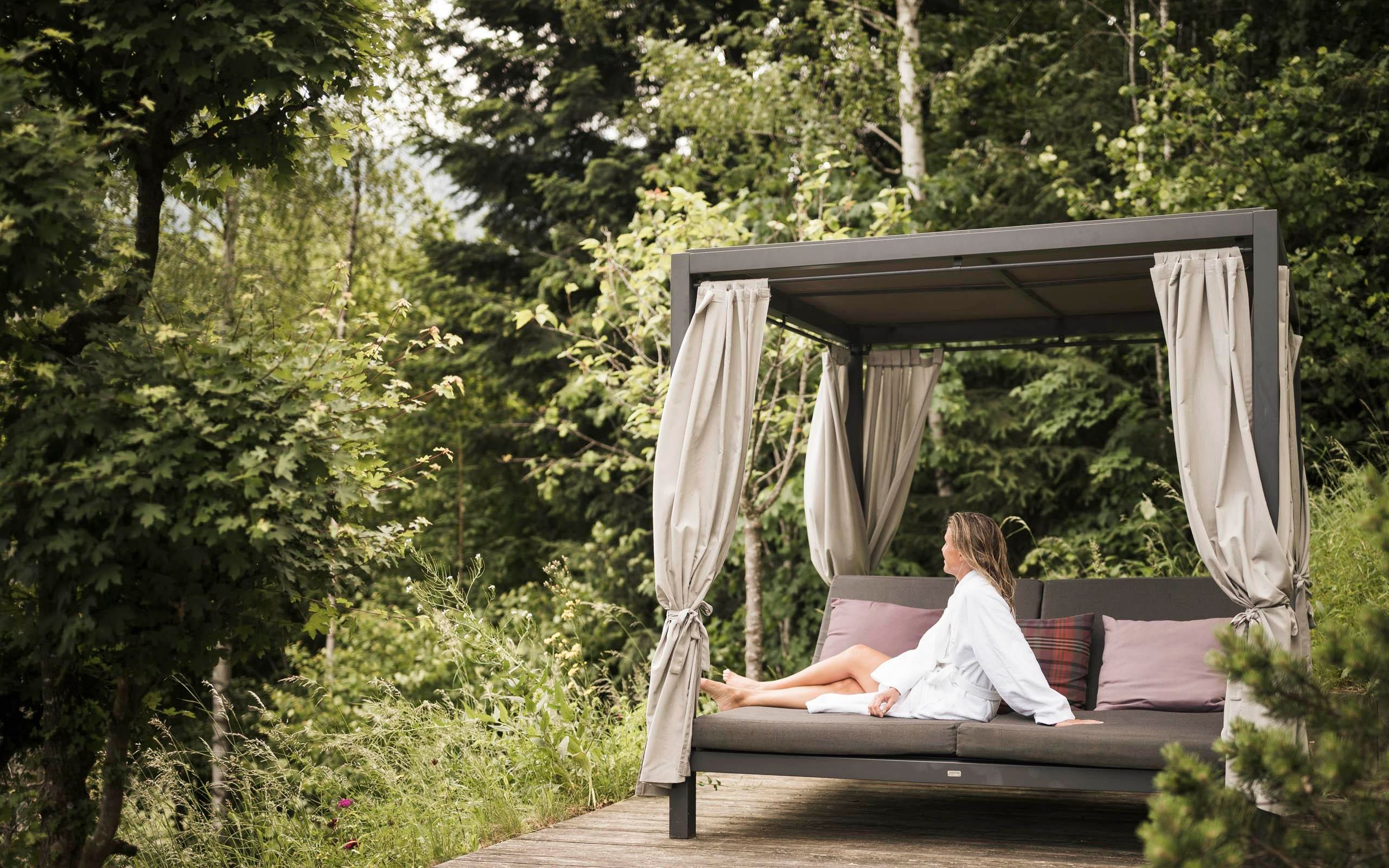 A woman sits comfortably in a cozy pavilion amidst green grass and trees. The place exudes calmness and a connection to nature.