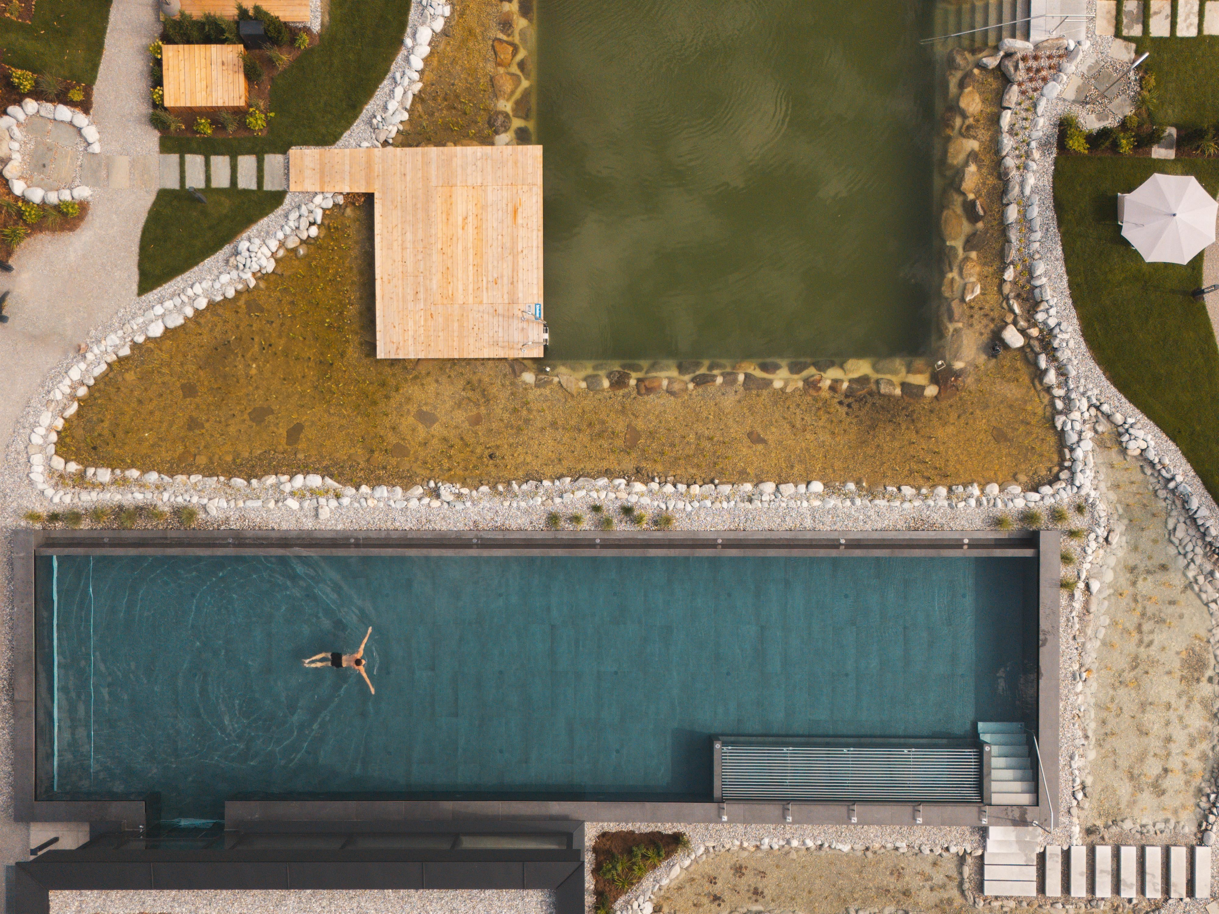 A beautiful pool in a green environment with a wooden deck. A person is swimming relaxed in the water.