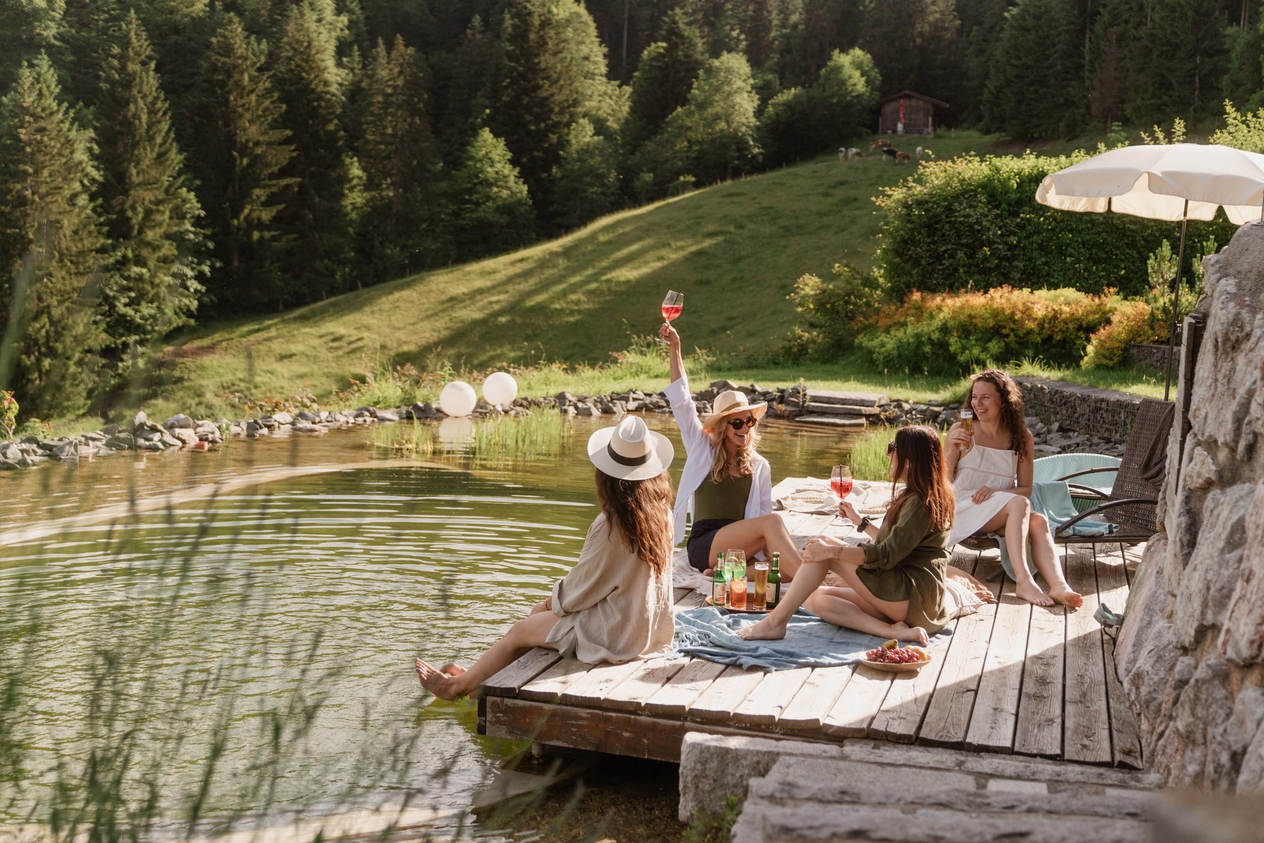 A group of women relaxes by the shore of a tranquil lake. They enjoy drinks and nature in a beautiful, sunny setting.