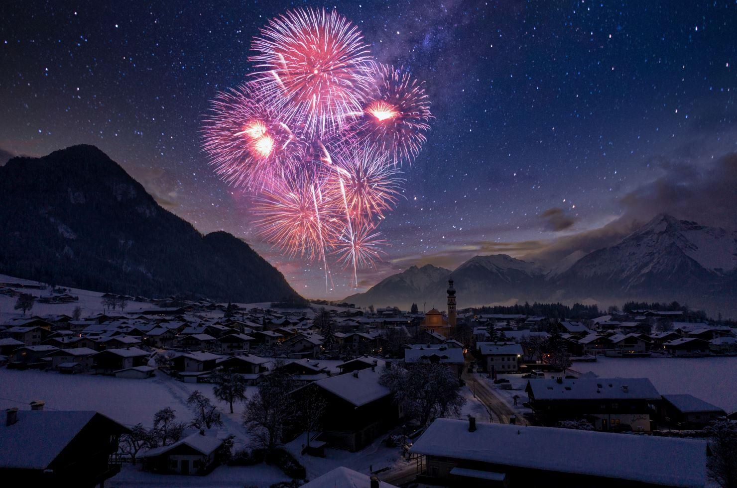 A breathtaking fireworks display lights up the night sky over a snow-covered village in the mountains. The landscape is calm and idyllic as the colorful lights illuminate the starry sky.