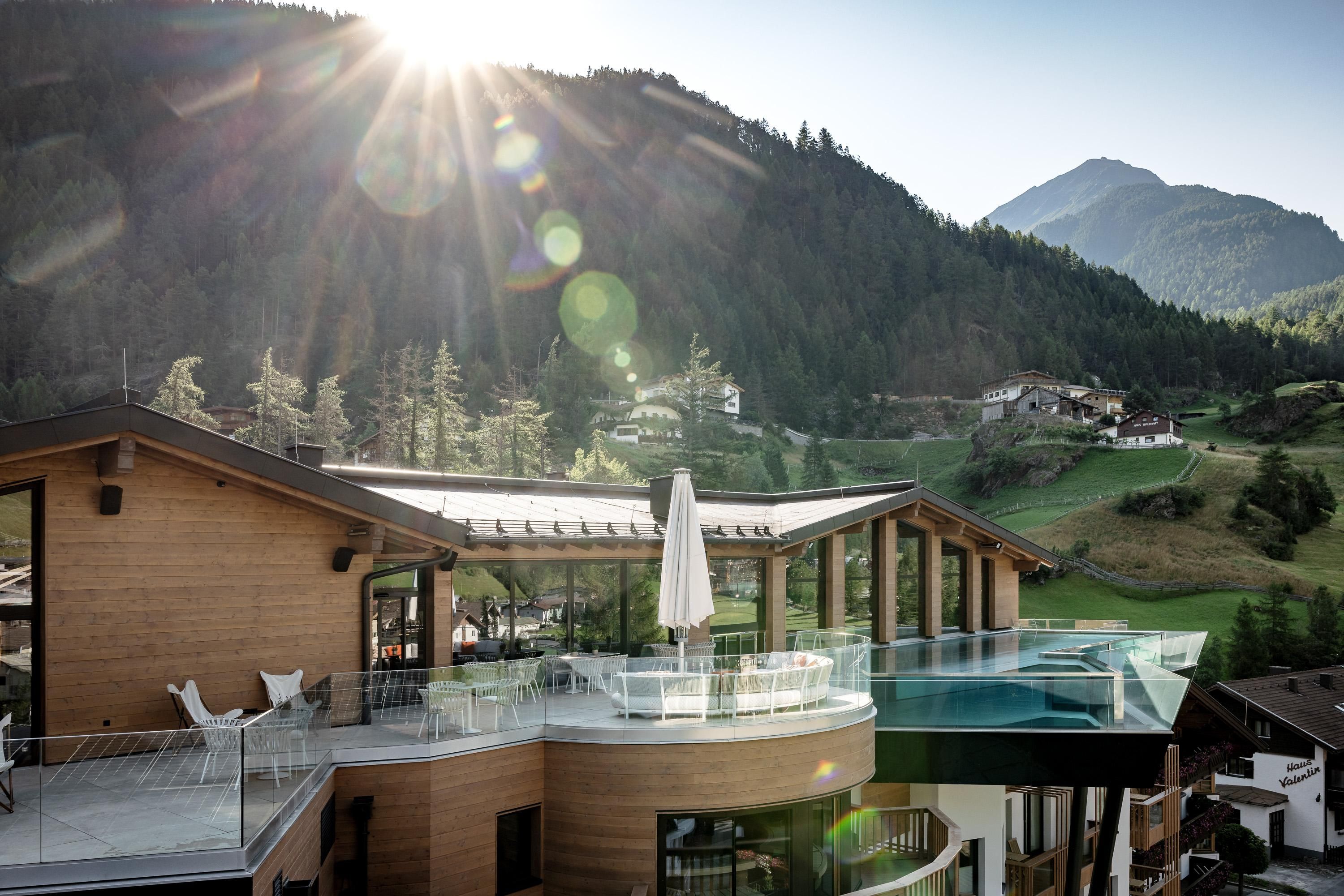 A modern building with a wooden facade and a large balcony. In the background, green mountains and a clear sky can be seen.