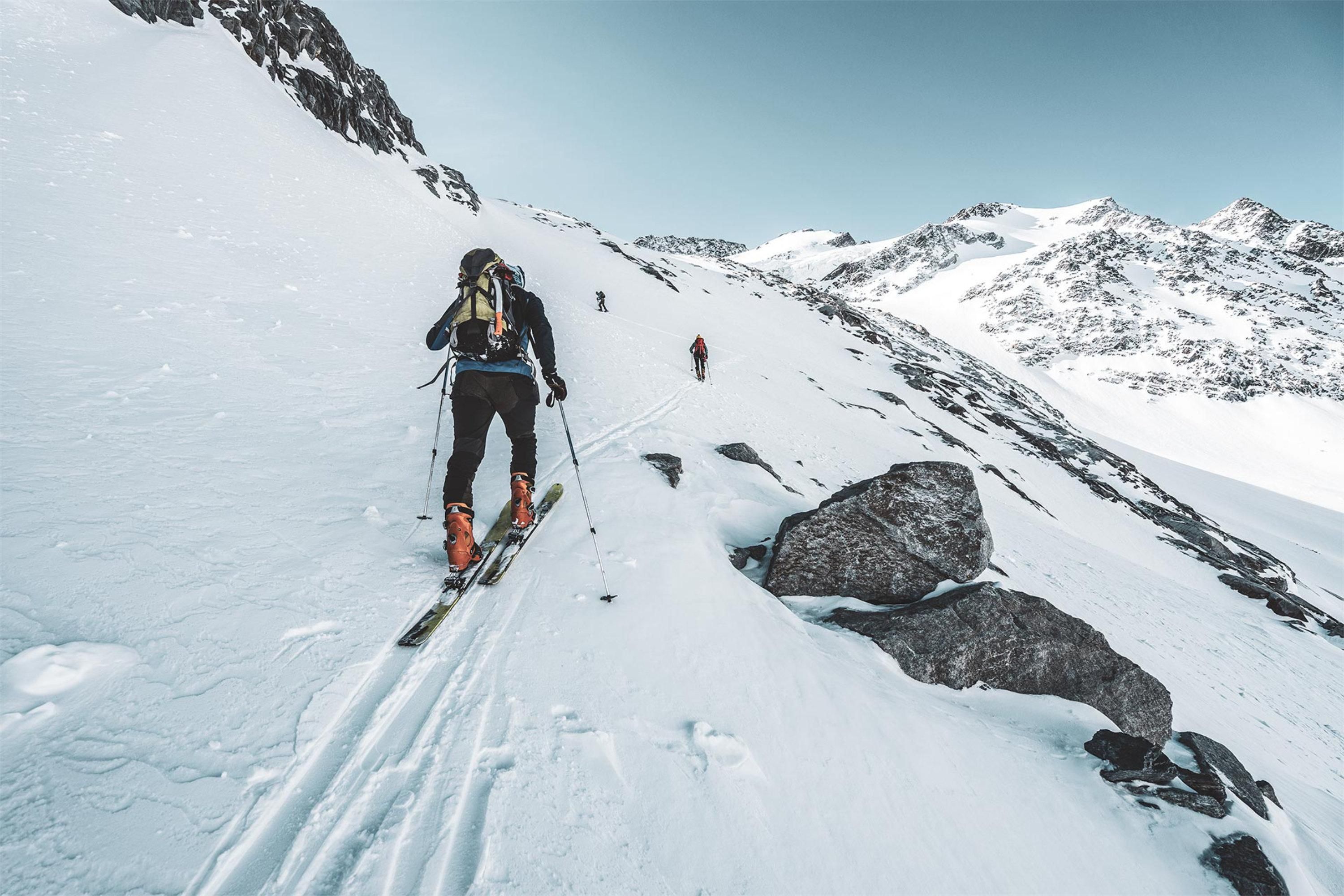 A skier climbs a snow-covered slope in the mountains. The surroundings are framed by impressive peaks and a clear sky.