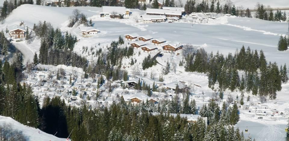 A winter landscape with snow-covered hills and many coniferous trees. Small, rustic cabins are integrated into the scene.
