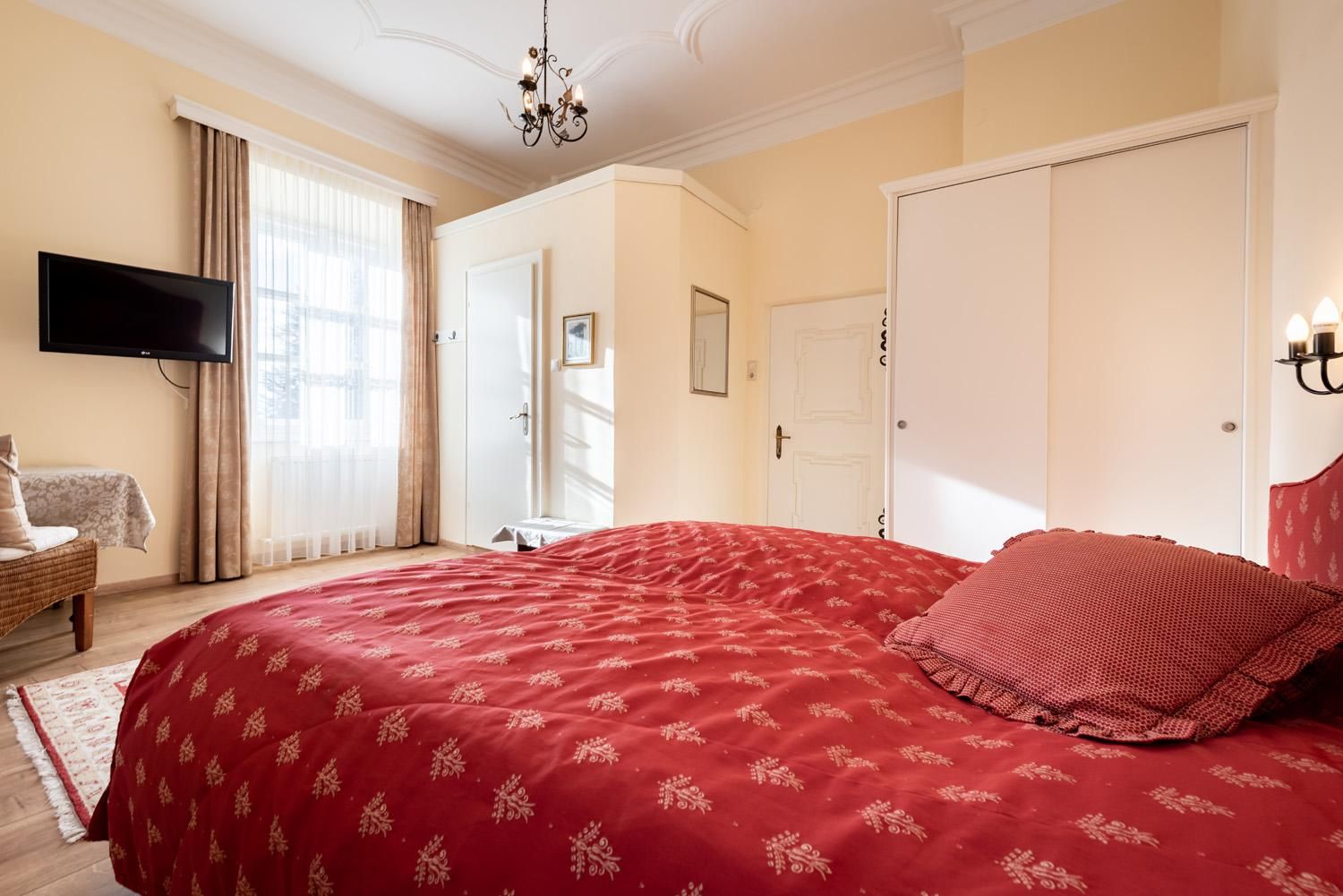 A cozy bedroom with a large red bed and bright walls. The room has a window front with curtains and a television on the wall.