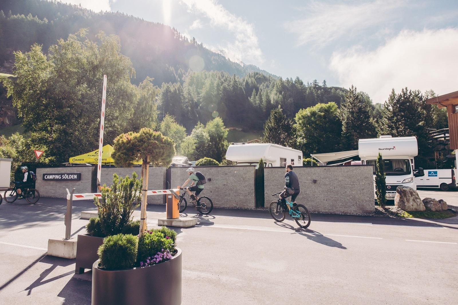 A beautiful day in a holiday resort with cyclists passing by an entrance. In the background, green trees and hills can be seen.
