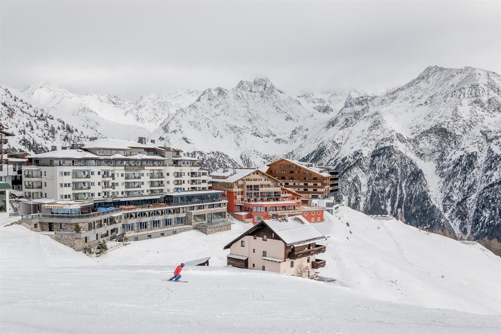 A snow-covered landscape with modern and traditional chalets. In the foreground, a skier is descending the slope, while the majestic mountains are visible in the background.