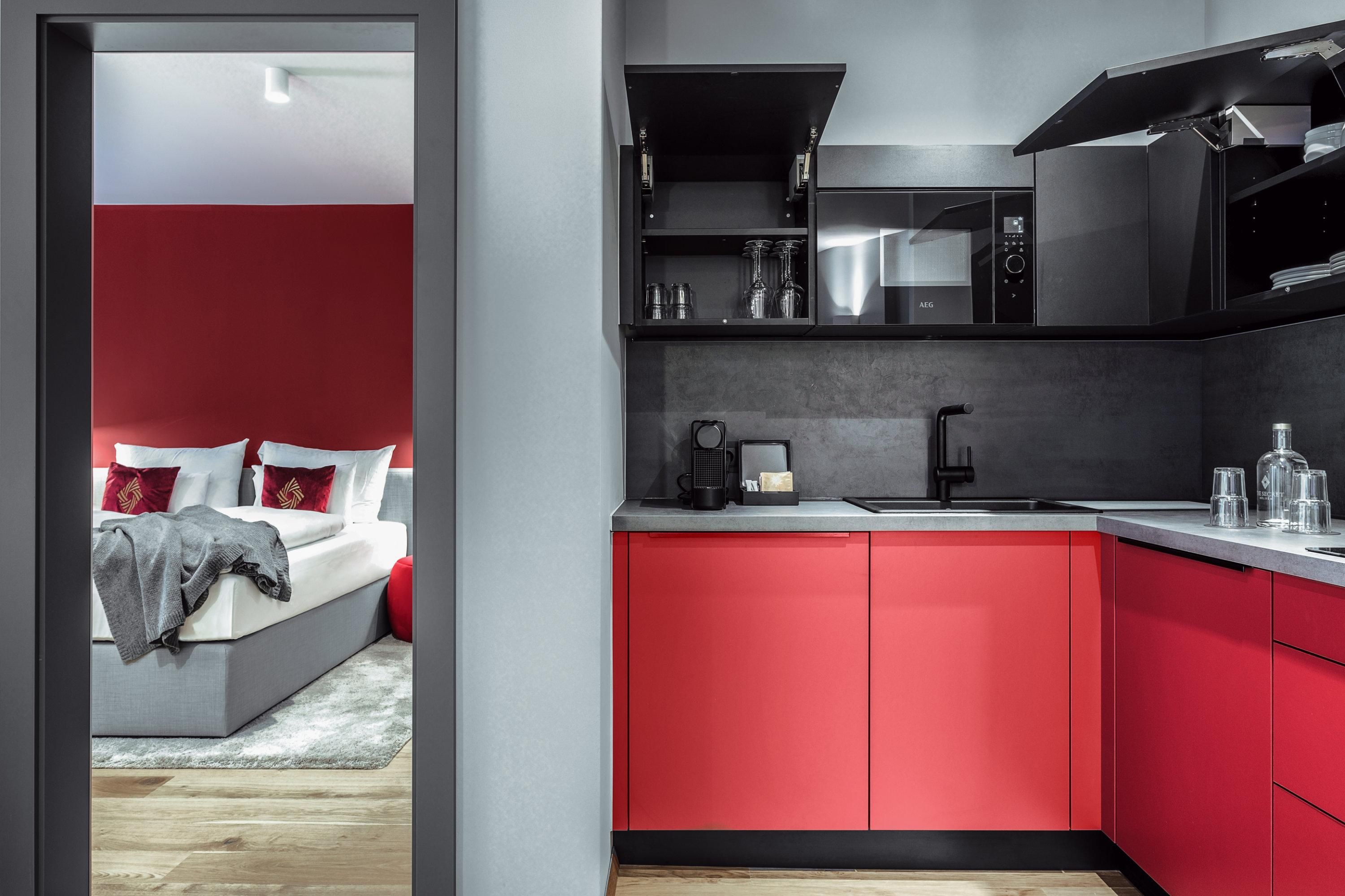 A modern kitchen with red cabinets and aclear work surface is visible. In the background, a cozy bedroom with white bedding and red accents can be seen.
