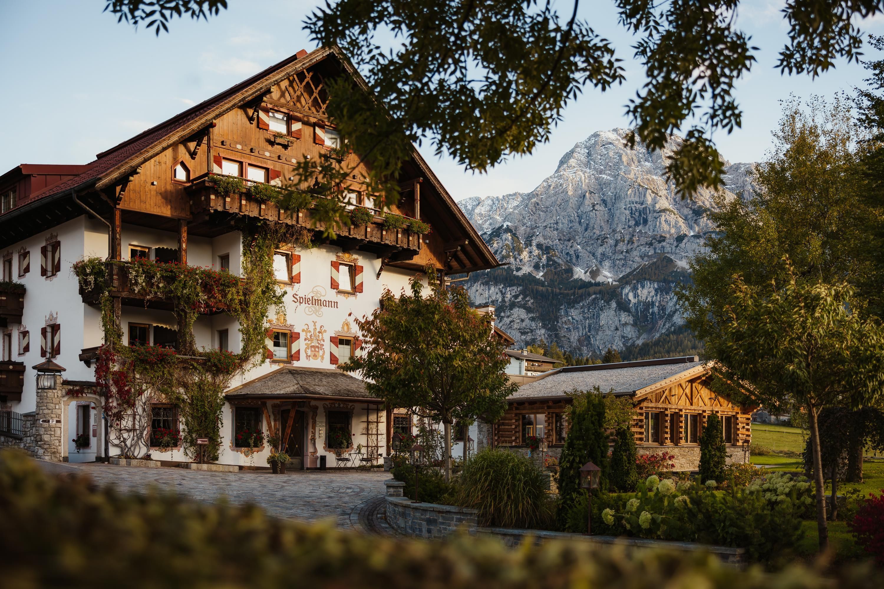 A charming farmhouse in the mountains, surrounded by trees and green meadows. The majestic mountains are visible in the background.
