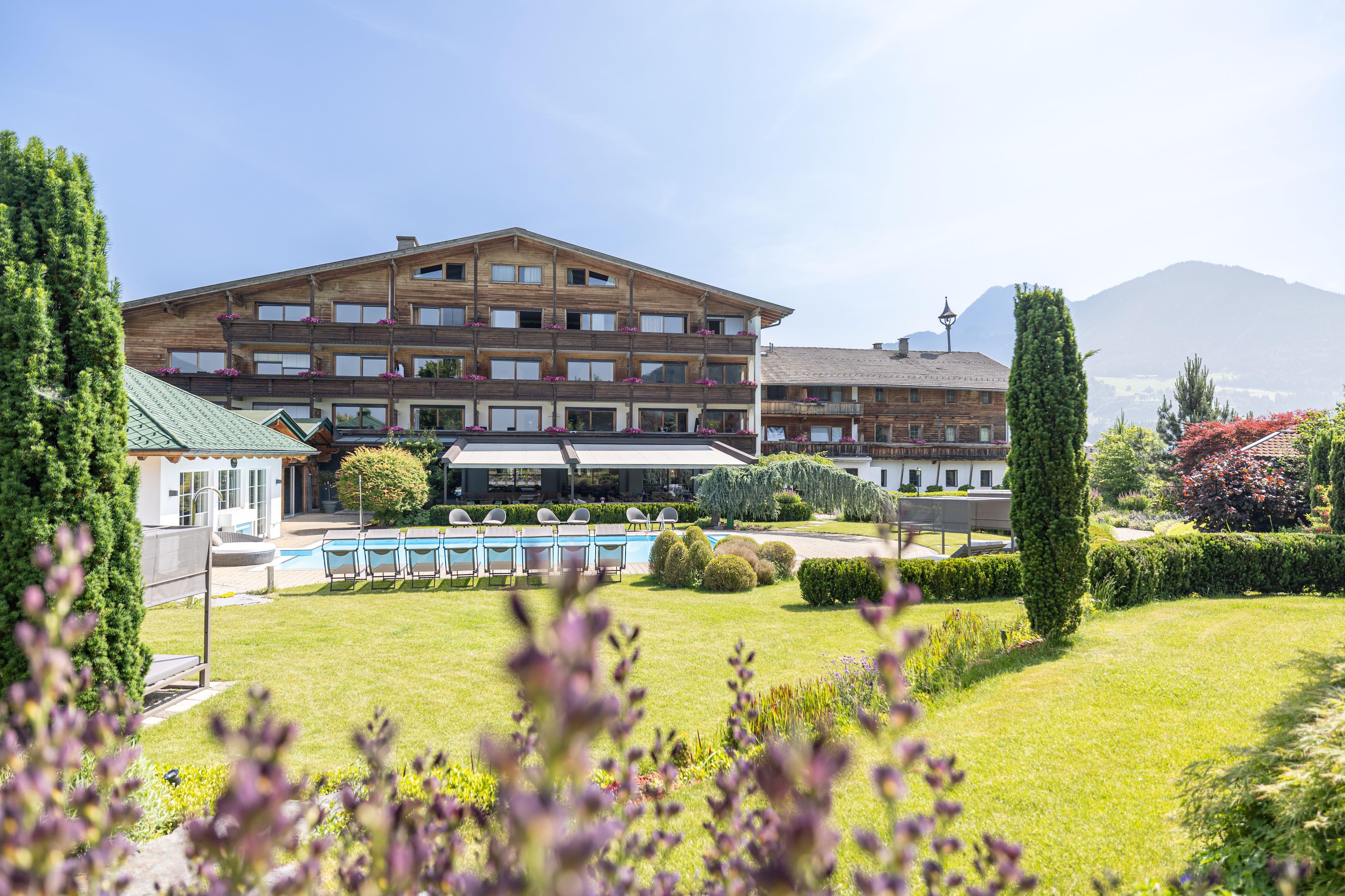 A beautiful hotel with a large garden and a pool. The mountains are visible in the background and the property is surrounded by green spaces.