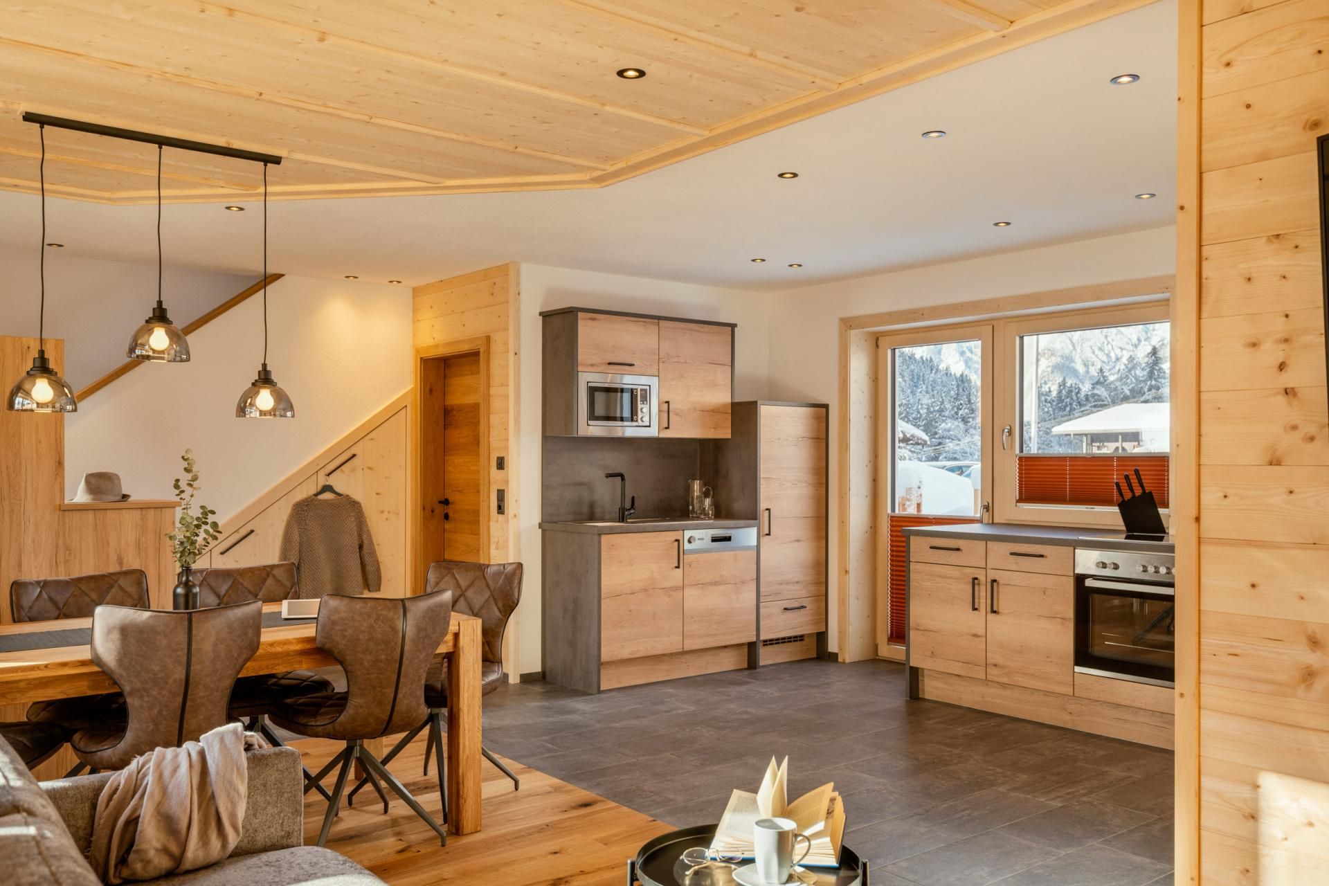 A modern kitchen with wooden cabinets and an open dining area. Large windows let in plenty of light and provide a view outside.