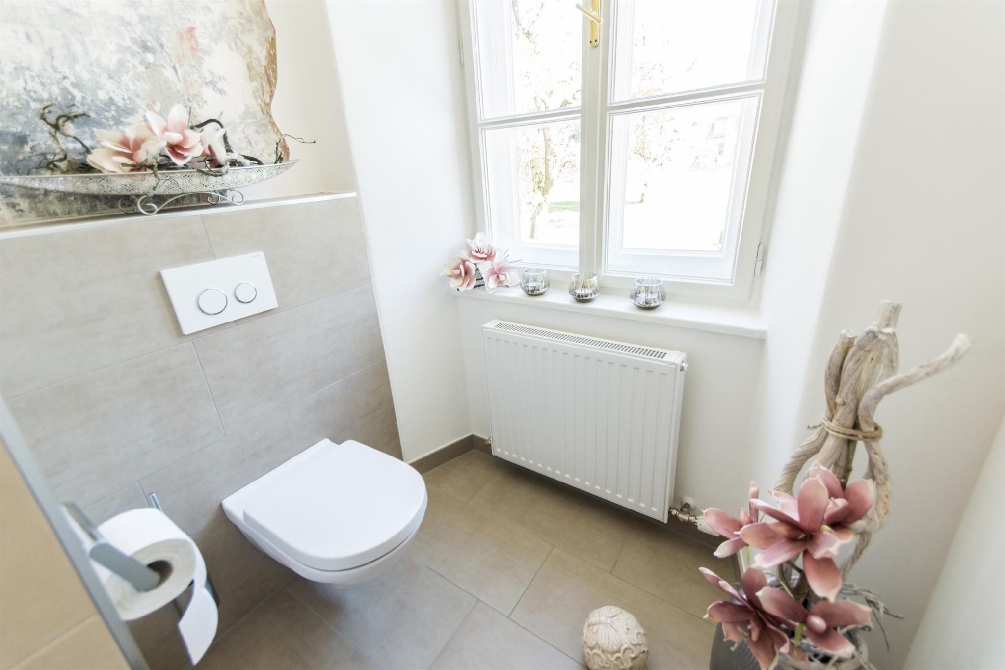 A modern bathroom with bright tiles and an elegant toilet. Next to the window is a flower arrangement and the heating is visible.