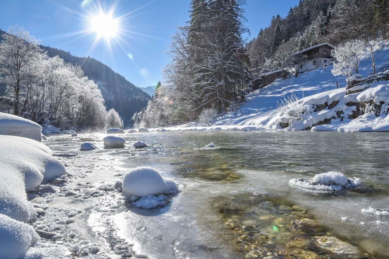 A winter landscape with a frozen river and snow-covered trees. The sun shines brightly in the sky and illuminates the scene.