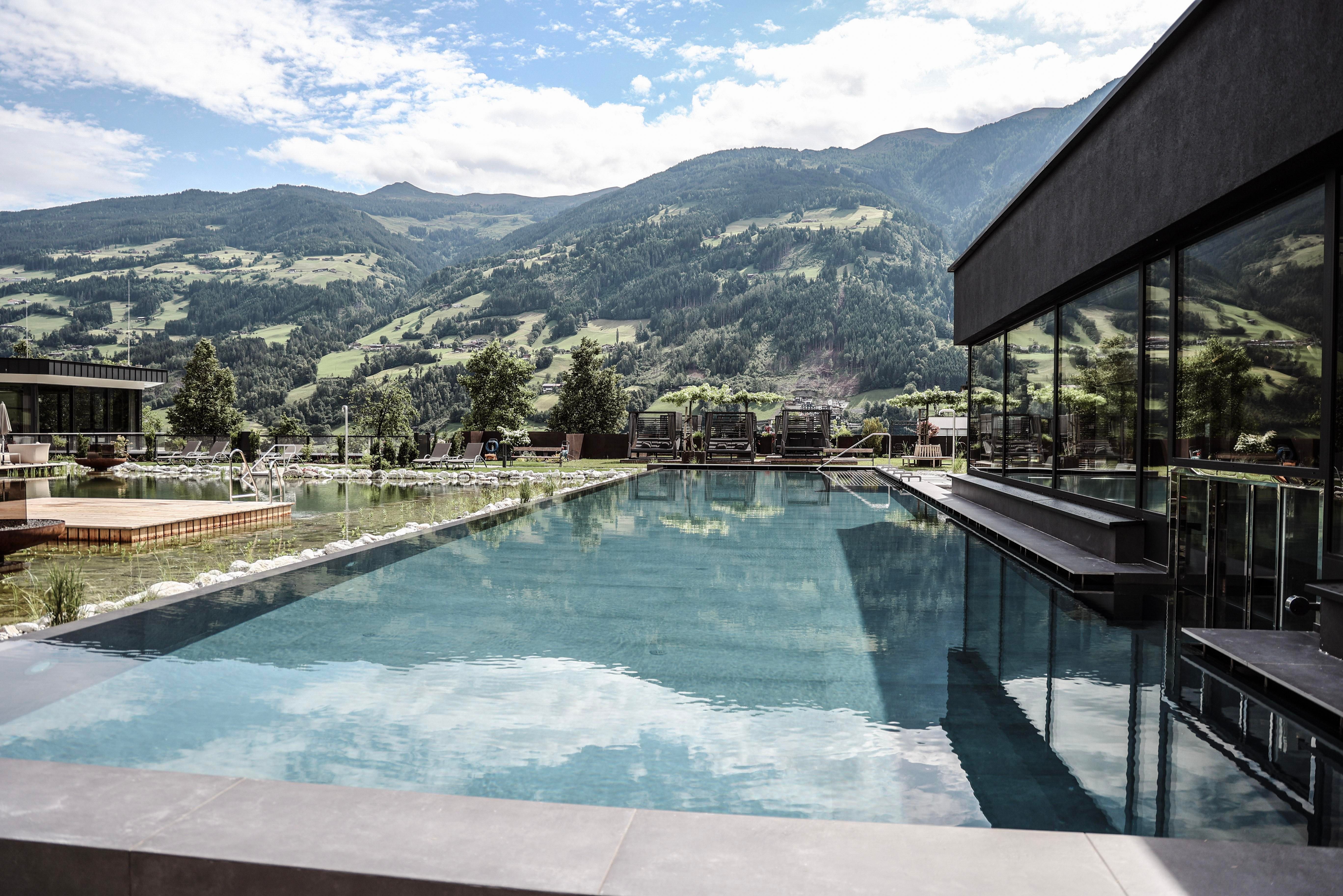 A modern infinity pool with clear water and a view of the mountains. The surroundings are green and scenic.