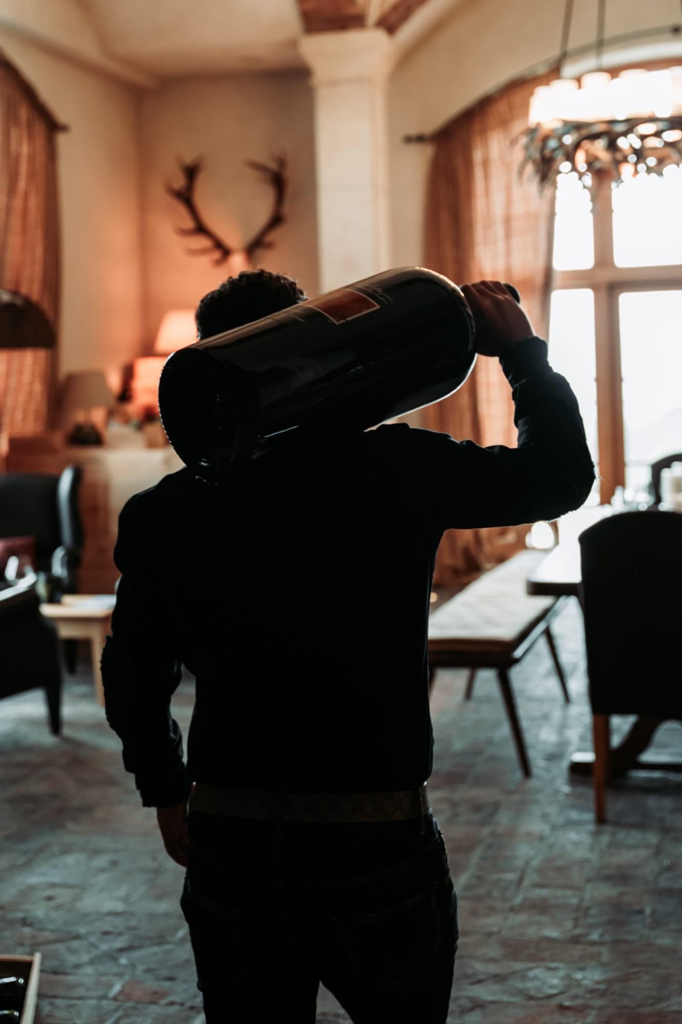A person with a large container on their shoulder stands in a rustic interior. The room is cozily furnished with wooden details and warm lighting.