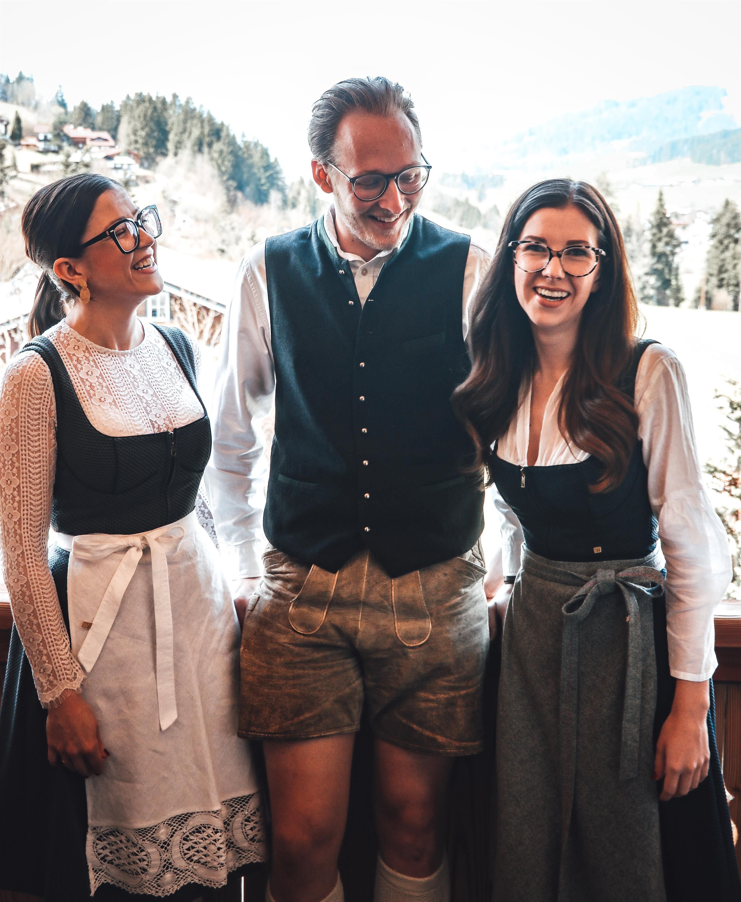 Three people in traditional Bavarian costume are standing in front of a wintry mountain landscape. They are smiling and appear friendly.