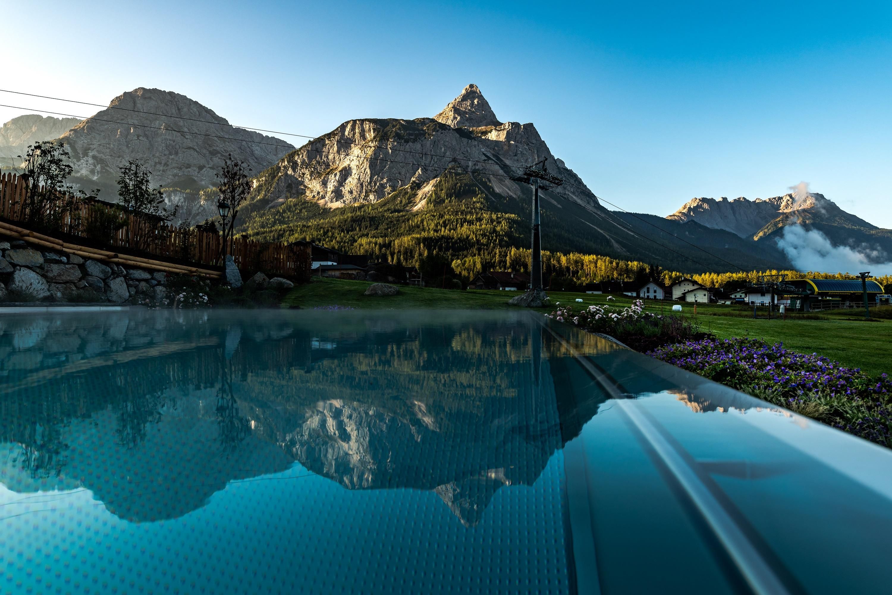 A tranquil pool with a clear water surface reflects the majestic mountains in the background. The landscape is surrounded by gentle hills and trees.