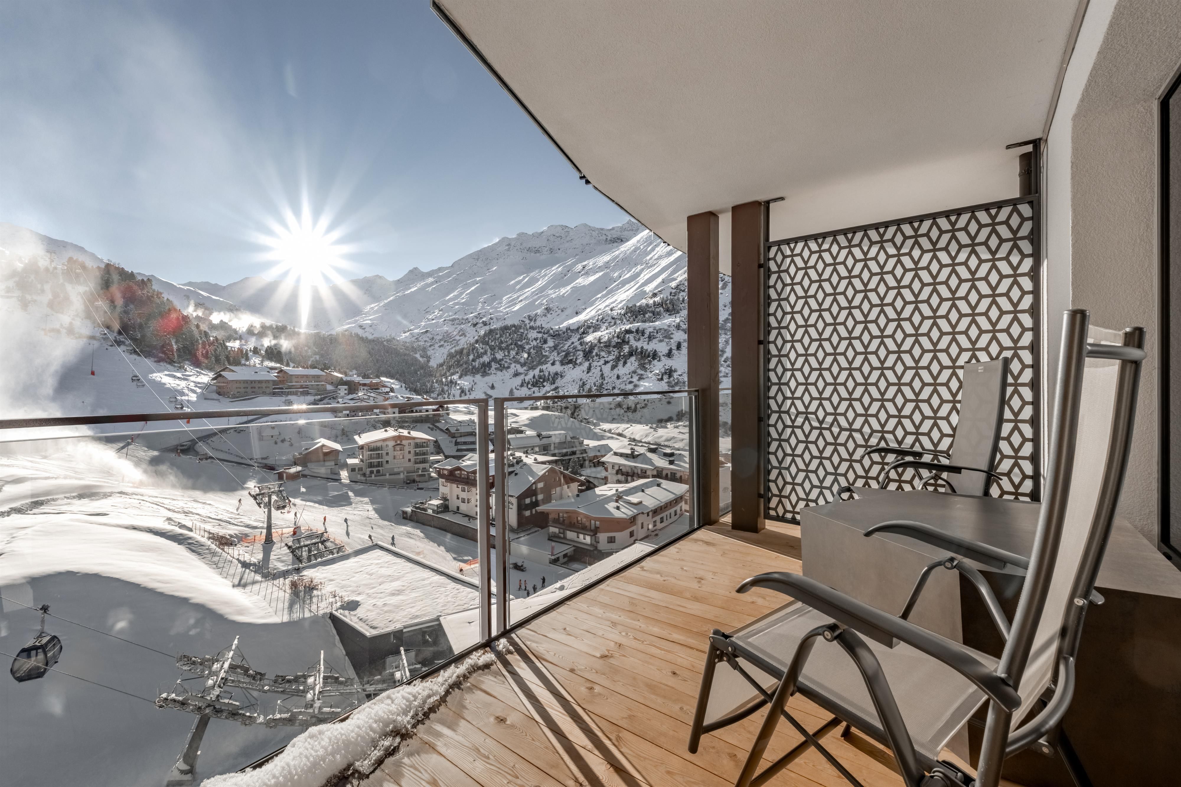 A balcony with a wooden floor and two chairs. In the background, snow-covered mountains extend and the sun is shining bright.
