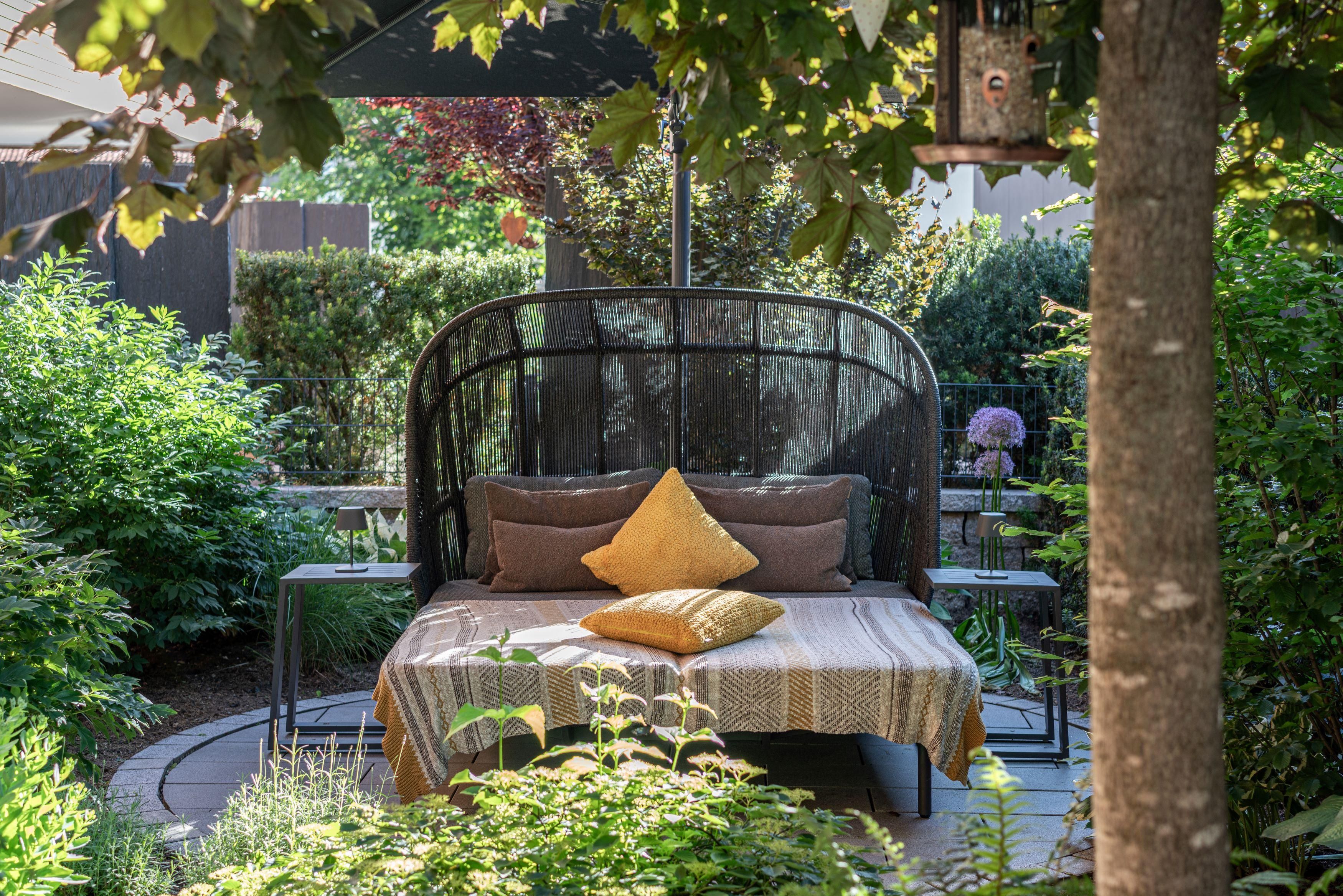A cozy seating area in the garden with a shaded lounge chair and colorful cushions. Surrounded by green plants and beautiful vegetation.