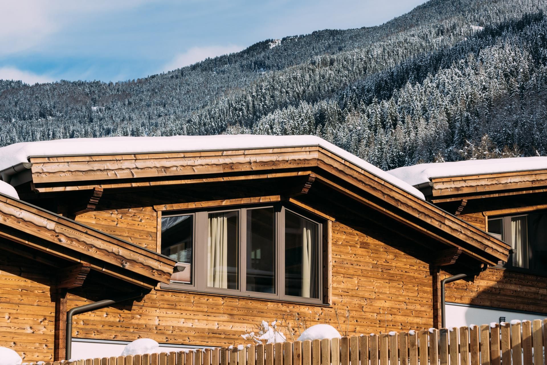 A modern wooden house with a snow-covered roof. Forested mountains can be seen in the background.