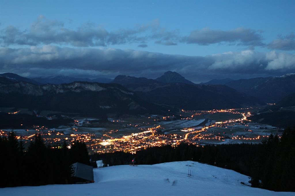 A quiet mountain landscape at night with snow-covered areas. The town in the valley glows softly and is surrounded by mountains.