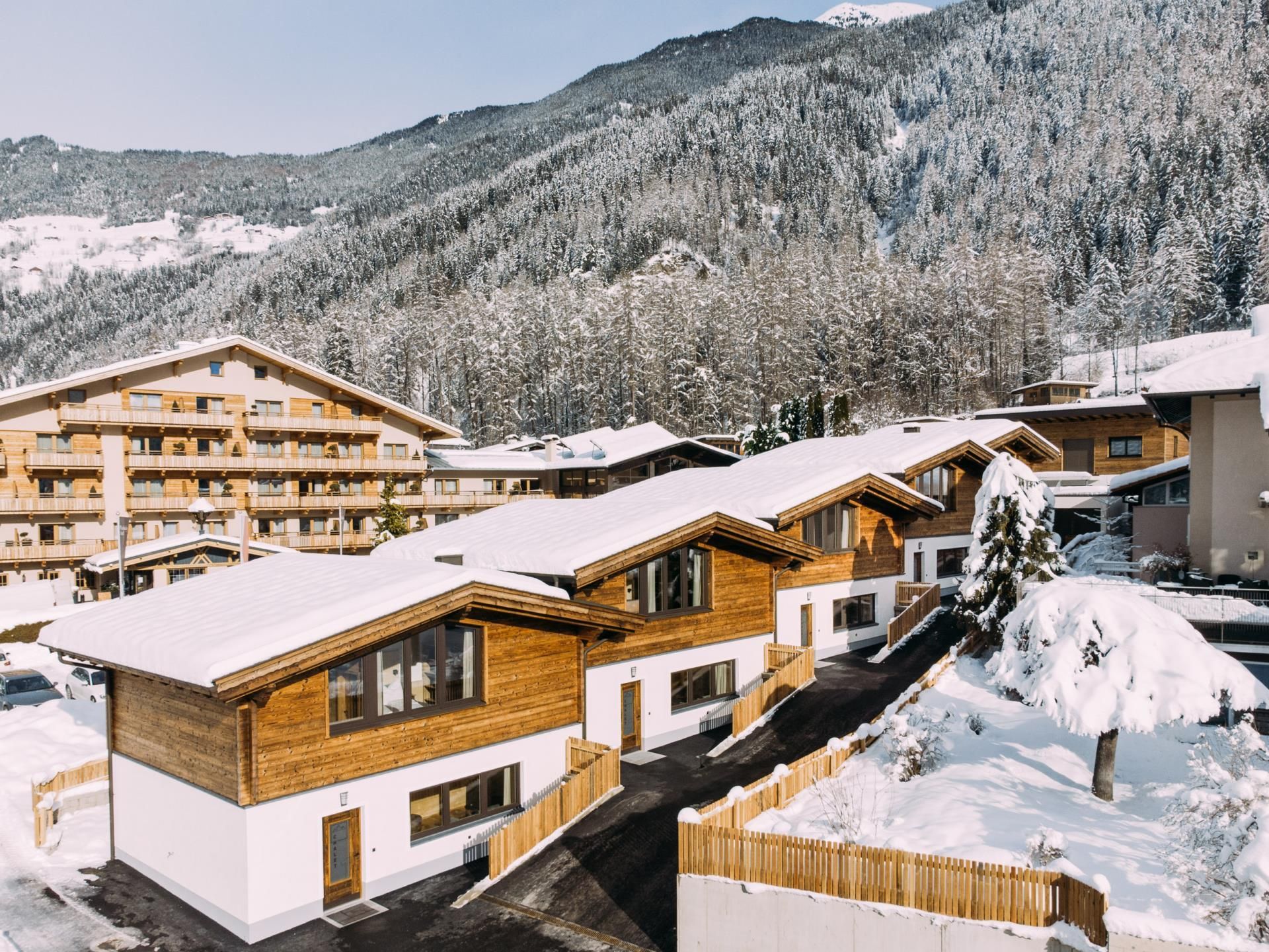 Modern holiday homes in a snowy landscape. In the background, high mountains and a clear sky can be seen.