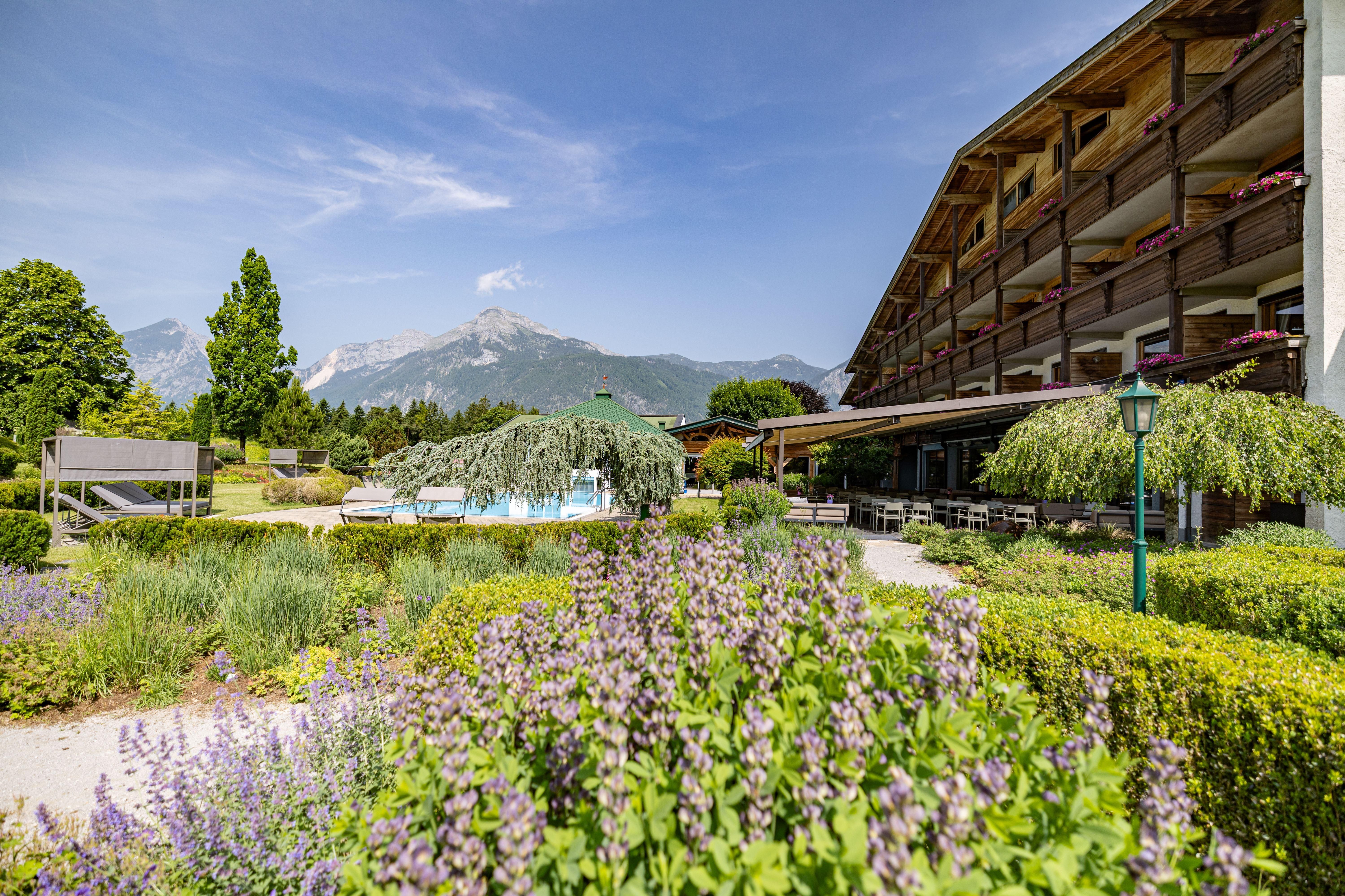 A picturesque garden with colorful flowers and a swimming pool. In the background, the mountains and a modern building can be seen.