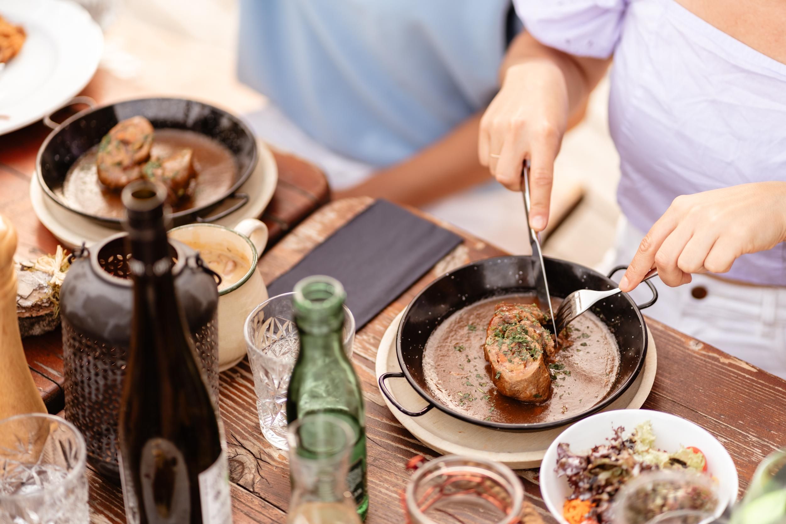 A set table with various dishes and drinks. One person is cutting a piece of meat on a plate.