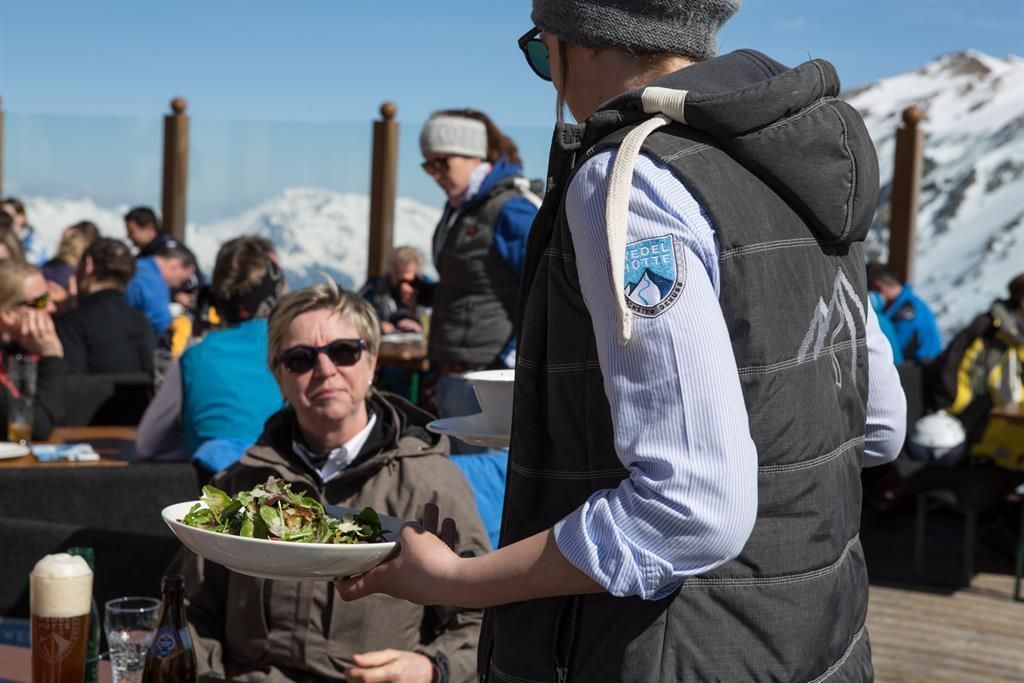 A person is serving a salad in a mountain restaurant setting. In the background, other guests and snow-covered mountains can be seen.