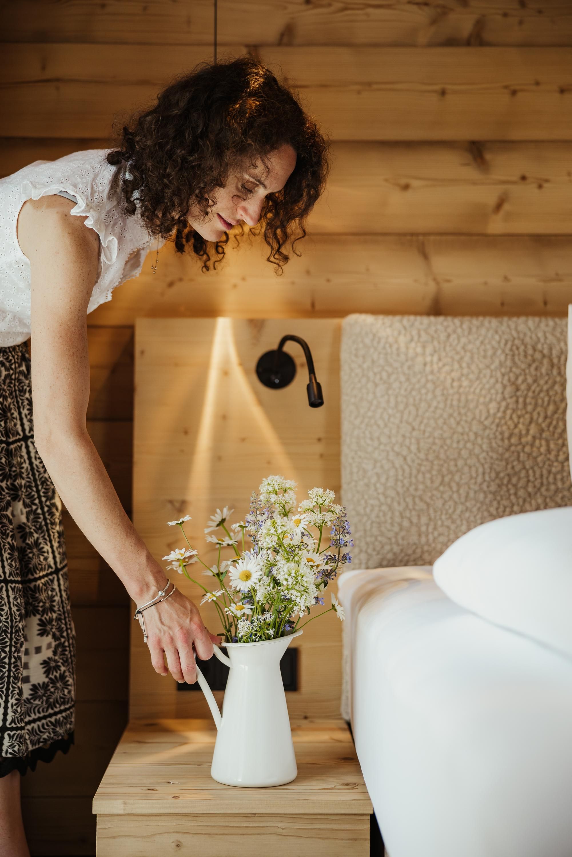 A woman places a bouquet of flowers in a white vase on a nightstand. In the background, a cozy room with wooden paneling can be seen.