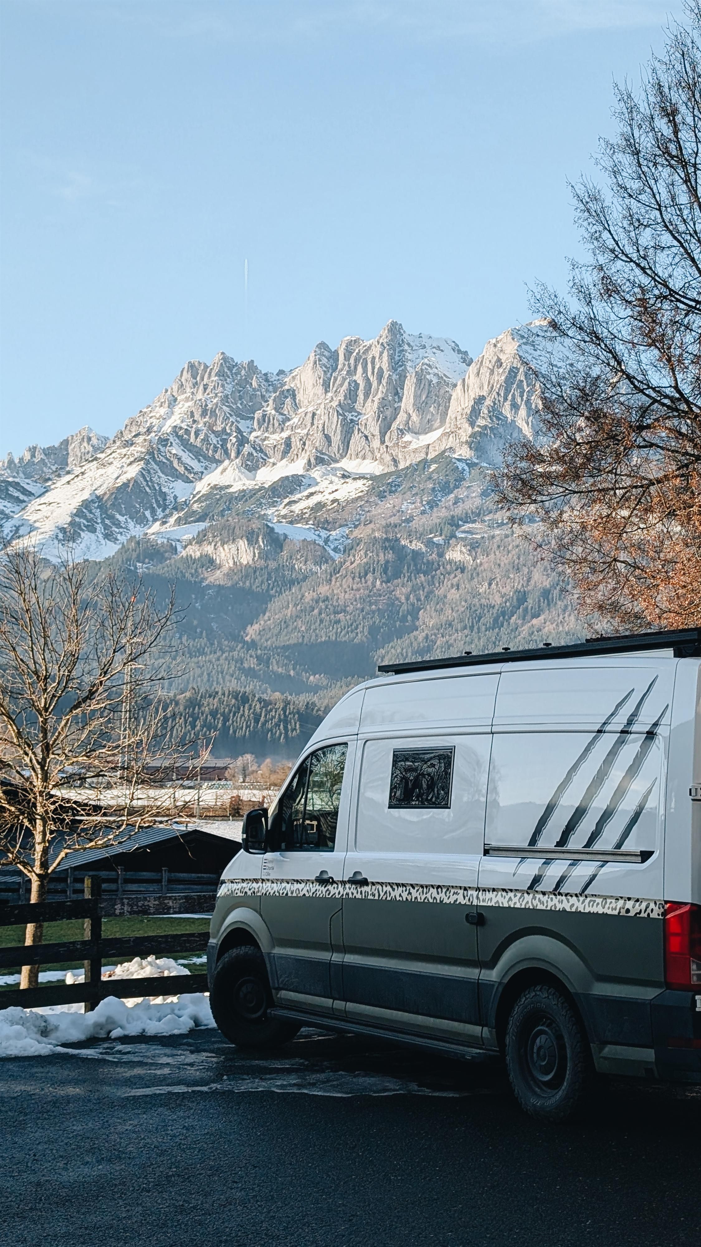 A motorhome is parked in a lot with a view of majestic mountains. The landscape is snow-covered and the sky is clear.
