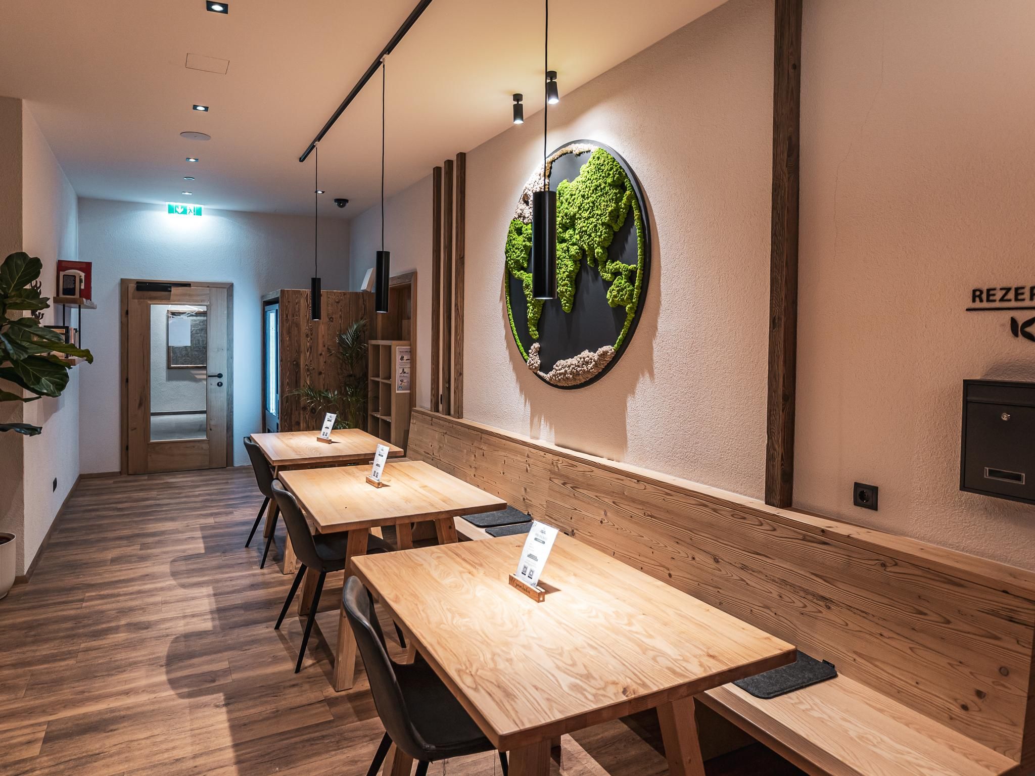 A modernly furnished room with wooden tables and chairs. A green wall decoration and soft lighting create an inviting atmosphere.