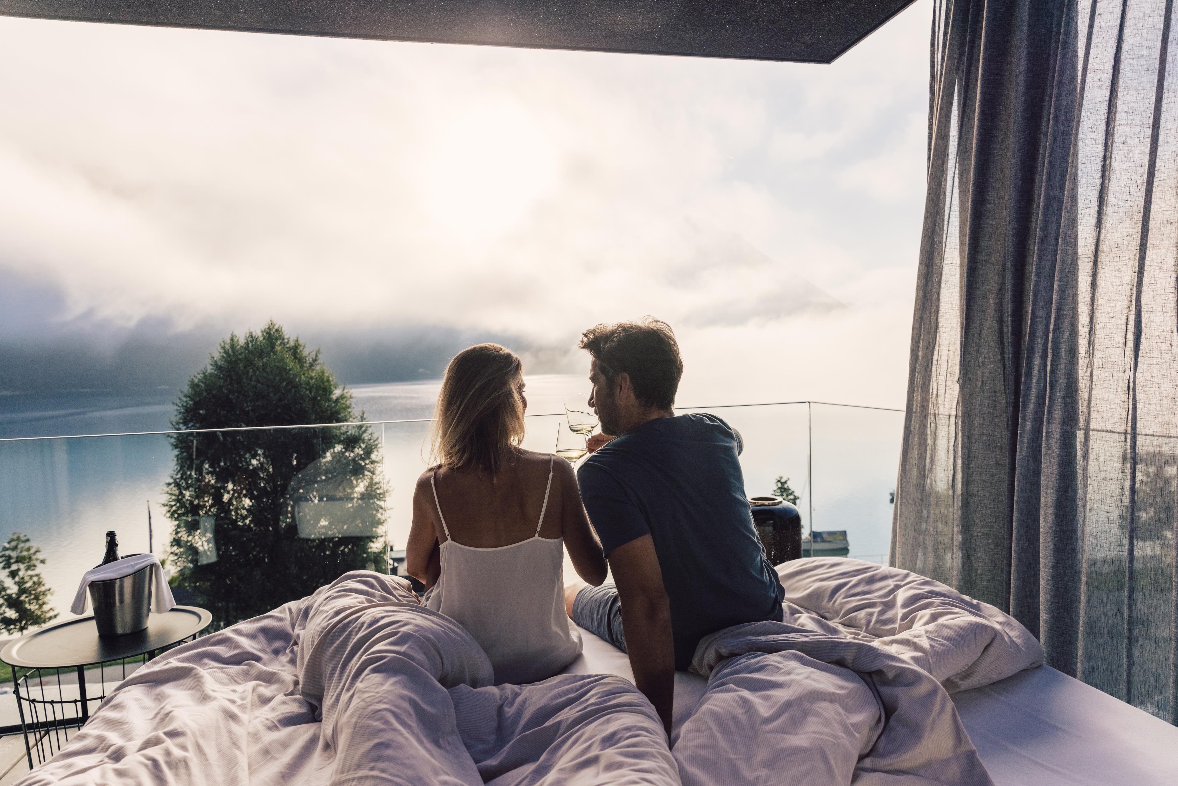 A couple is sitting on a bed outdoors, looking at a picturesque landscape. The atmosphere is romantic and relaxed, with a view of water and mountains.