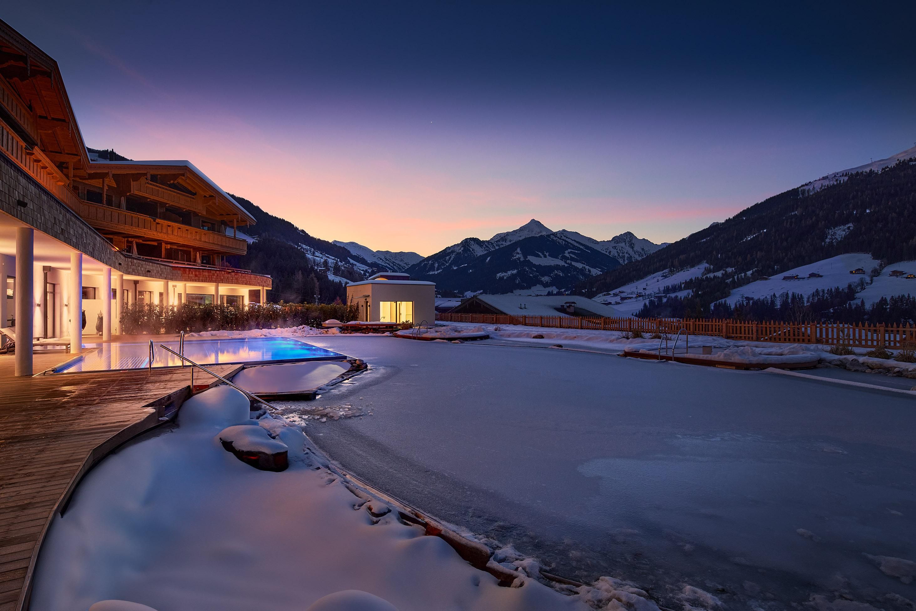A quiet winter evening with snow-covered mountains and a frozen lake. The architecture integrates harmoniously into the mountain landscape.