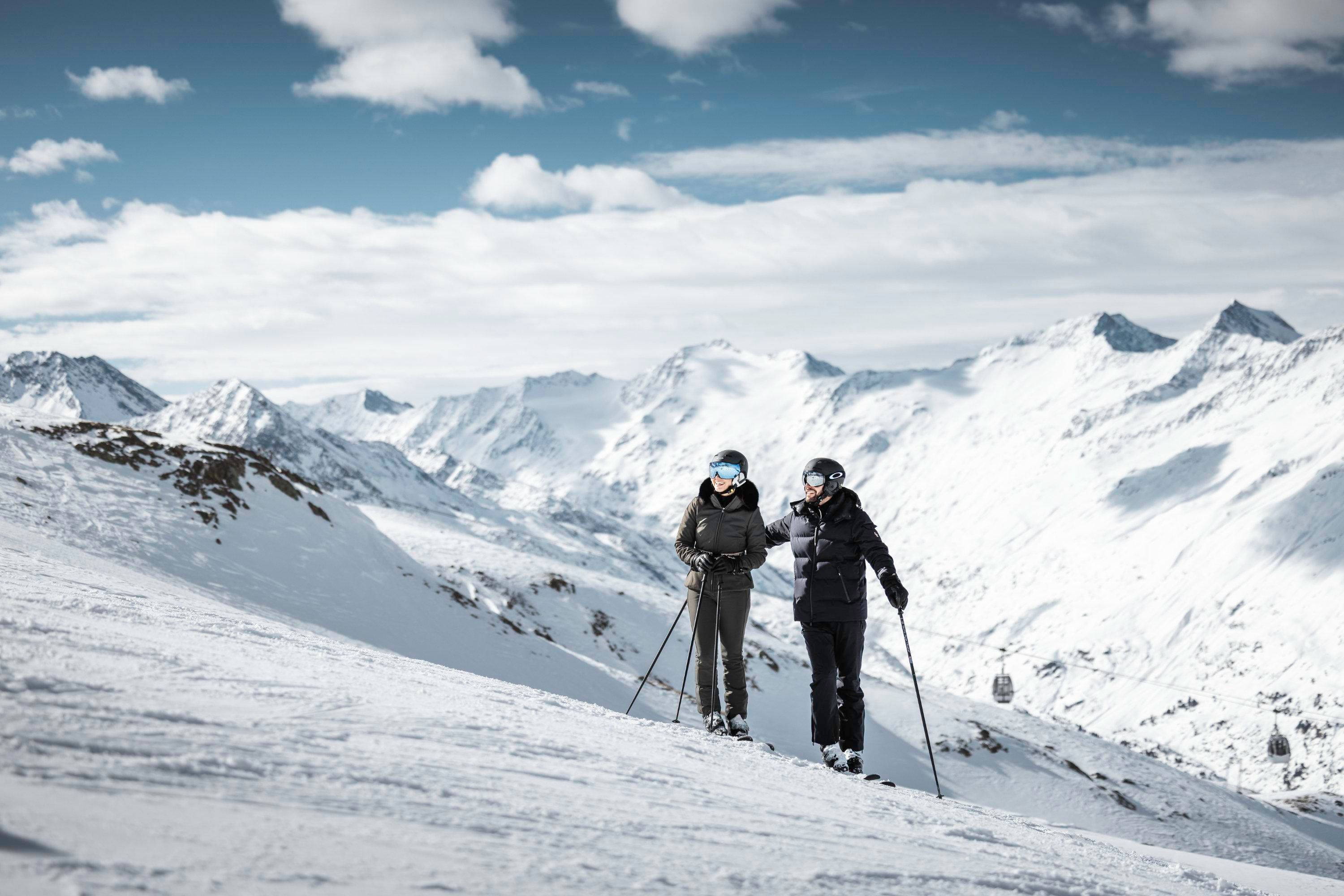 Two people are standing on a snow-covered slope in the mountains. The sky is clear with some clouds, and the surroundings are wintry and beautiful.