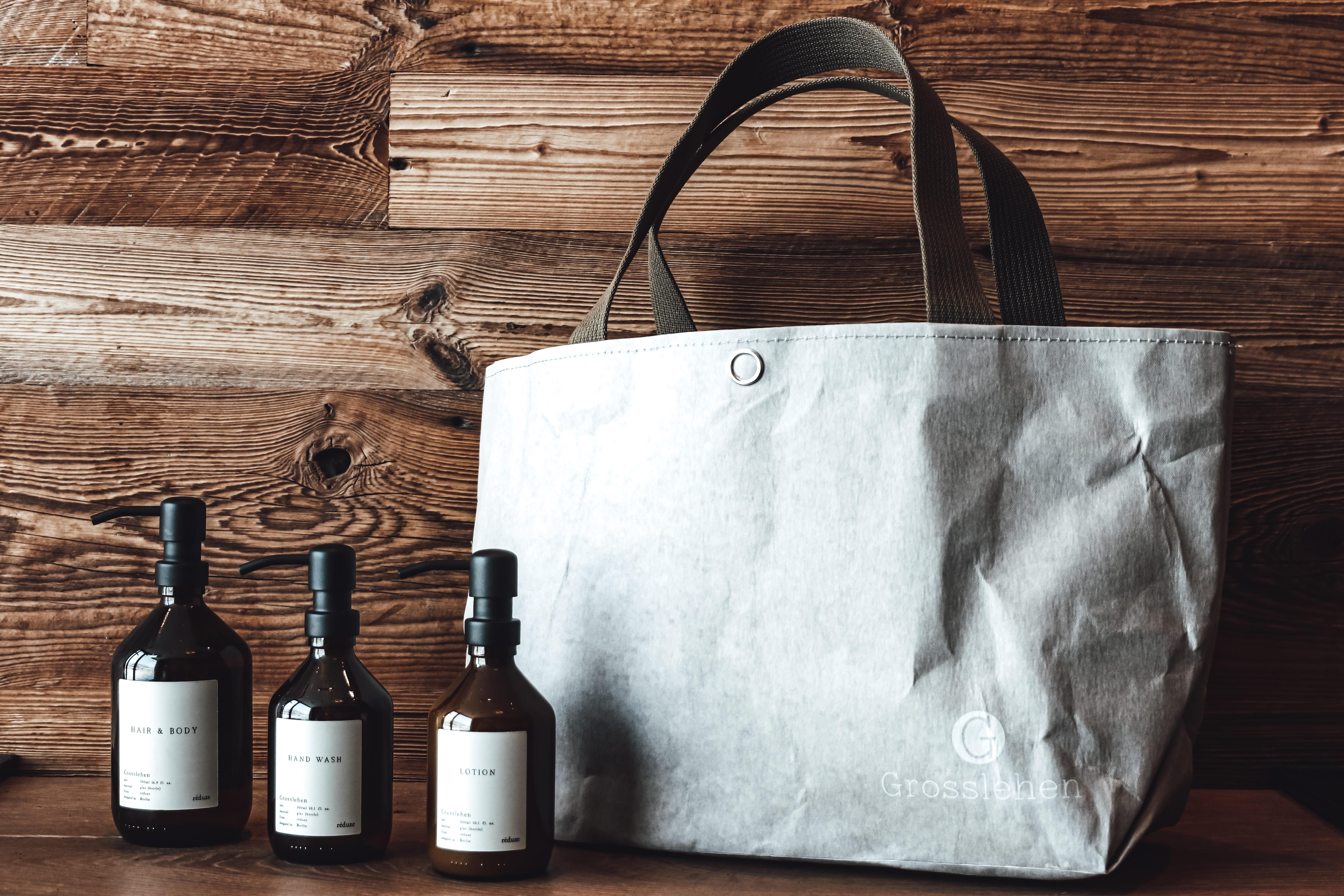 A stylish bag made of gray material stands next to three elegant bottles on a wooden table. The bottles have simple labels and complement the modern, minimalist design.